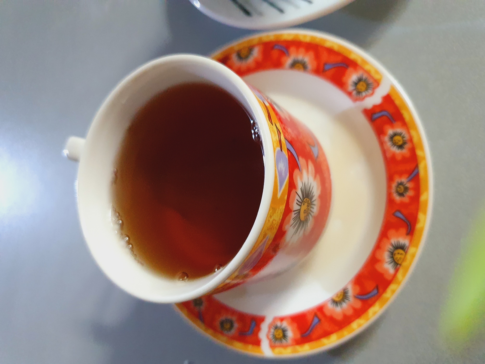 A close-up, overhead shot shows a cup of tea and its saucer. The cup is white with a brightly colored floral pattern of orange, yellow, and purple flowers. The saucer shares the same floral design. The tea is a dark amber color with a few small bubbles on the surface. The image appears to be taken indoors, on a reflective surface that suggests a tabletop. There are no people or animals visible in the image. The setting does not provide any specific indicators of the city of Shiraz, Iran.