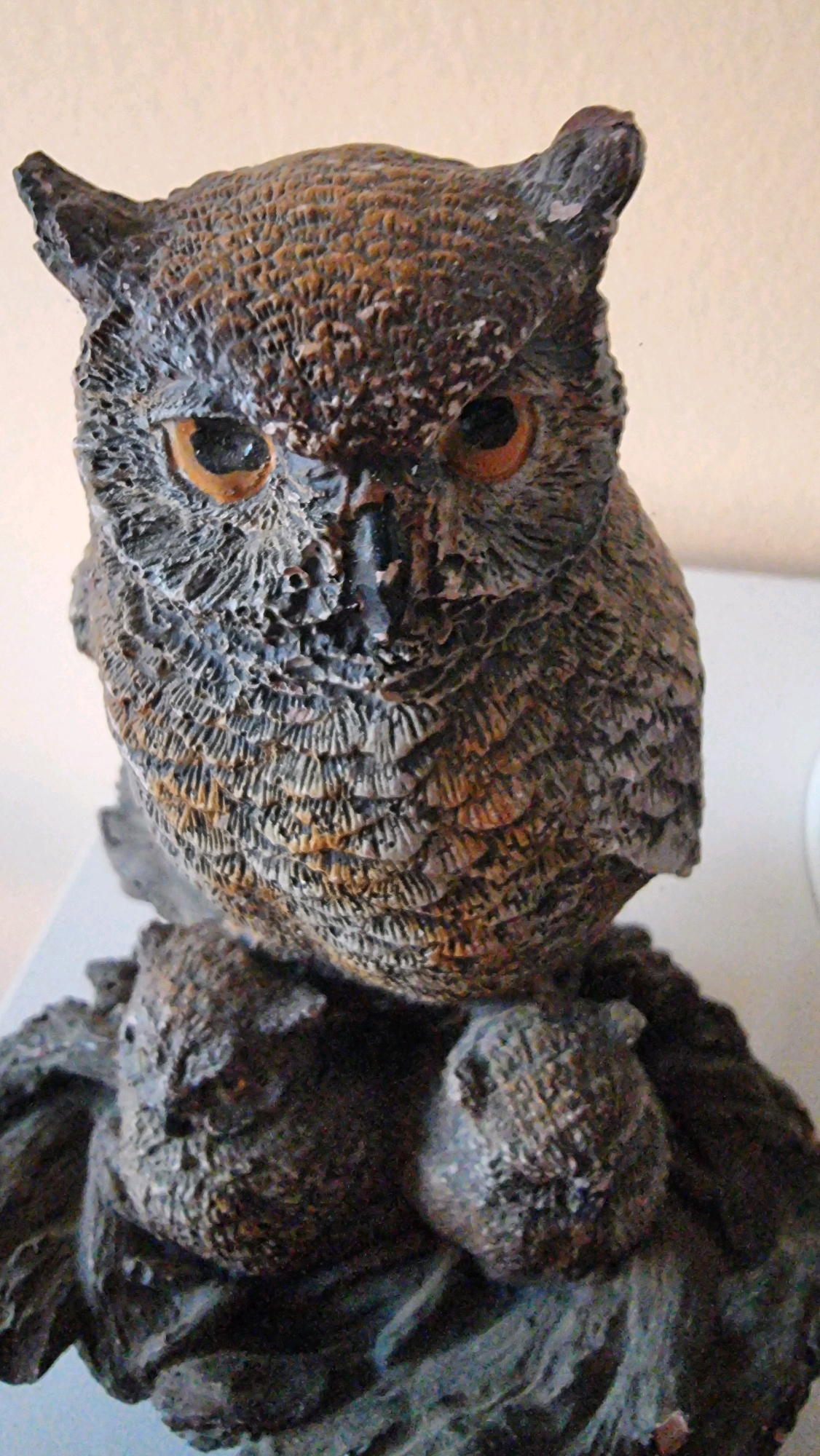 A close-up, head-on, vertical shot features a detailed owl figurine perched on a rocky base. The figurine is made of dark brown and grey material with textured detailing to mimic feathers. The owl's eyes are round and orange, and it has prominent ear tufts. The base appears to be made of dark, rough material resembling rocks or bark, with some lighter grey and brown areas. The background is a plain, light beige wall. The image does not depict the city of Sant Joan Les Fonts, Spain, nor does it contain any people, actions, or interactions.