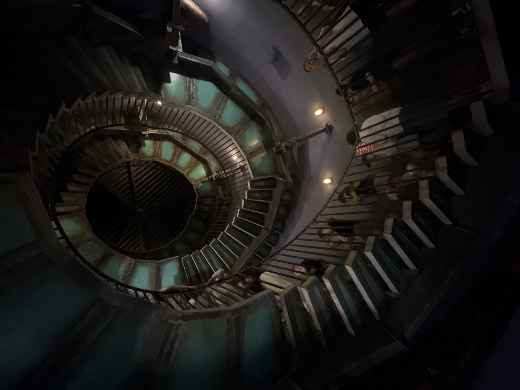 The image shows a low-angle view of a spiral staircase in a dark environment. The staircase is constructed of metal and concrete, with a teal-colored accent on the risers. The walls surrounding the staircase are painted gray and lit by dim yellow lights. People are visible on the upper levels of the staircase, though they are somewhat obscured by the low lighting. The scene depicts a descending, winding path within an enclosed structure. This is the Revolution exhibition in Moscow, Russia, where visitors walk up a circular staircase.