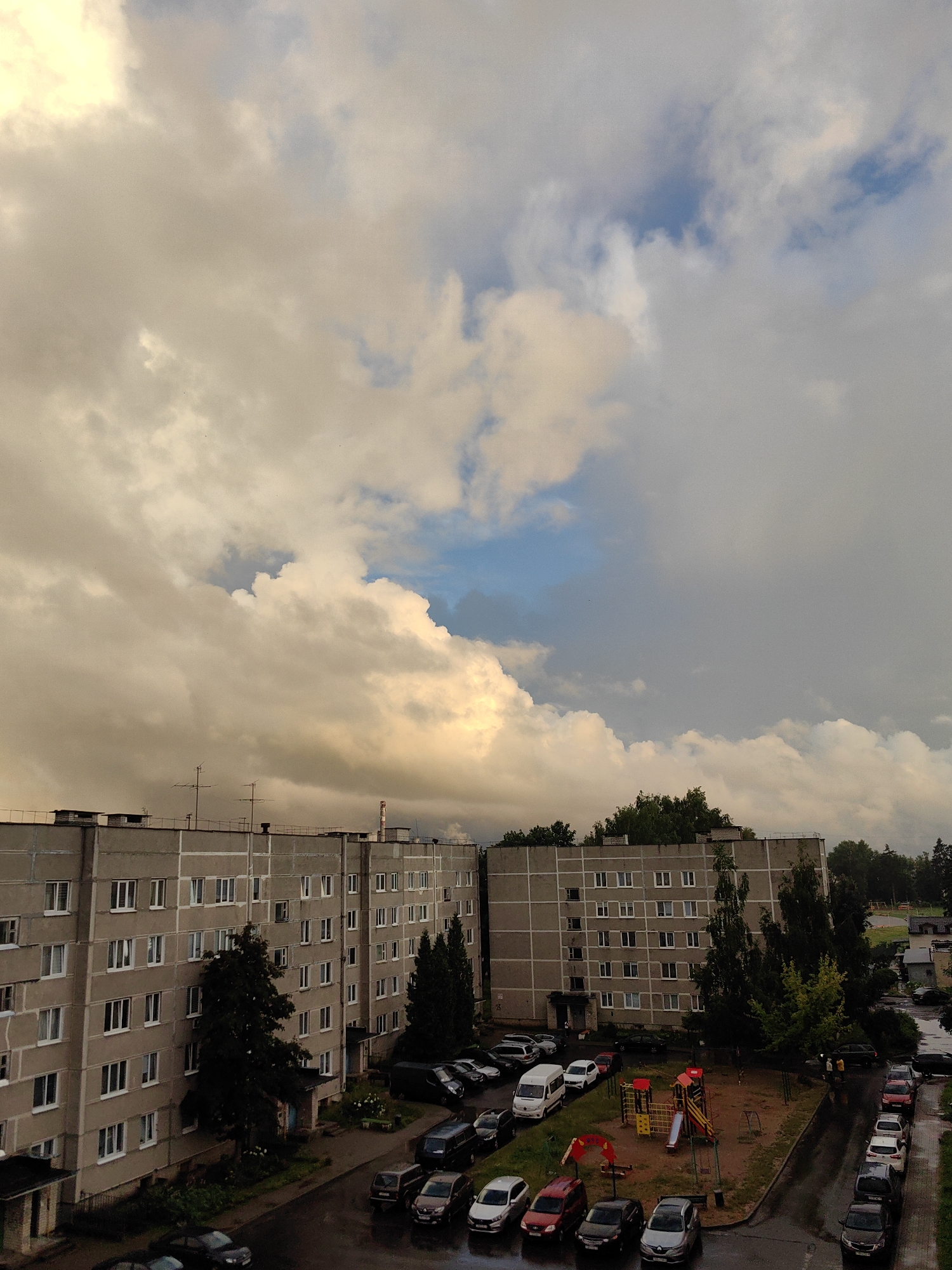 An aerial, eye-level shot captures a residential area in Fanipal, Belarus, under a dramatic sky filled with large, fluffy clouds tinged with yellow light and patches of blue. In the foreground, two identical, grey, five-story apartment buildings face each other across a wet, asphalt-paved courtyard. The buildings are rectangular with numerous windows, each divided into sections by white vertical and horizontal lines. An antenna array is visible on the roof of the left building, and a brick chimney with a red cap stands on the roof of the right building.
In the courtyard between the buildings, a playground with red and yellow structures, including a slide and swings, is set up on a dirt and gravel surface. Several cars are parked on the paved areas and along the edges of the courtyard. The vehicles are a mix of sedans, SUVs, and a white van, with colors ranging from black and grey to silver and red. Two people, appearing as small figures, are walking near the playground. Lush green trees and bushes are scattered around the buildings and line the perimeter of the scene, with some trees growing tall enough to reach the upper stories of the apartments. The overall atmosphere is somewhat overcast, with the wet pavement suggesting recent rain.