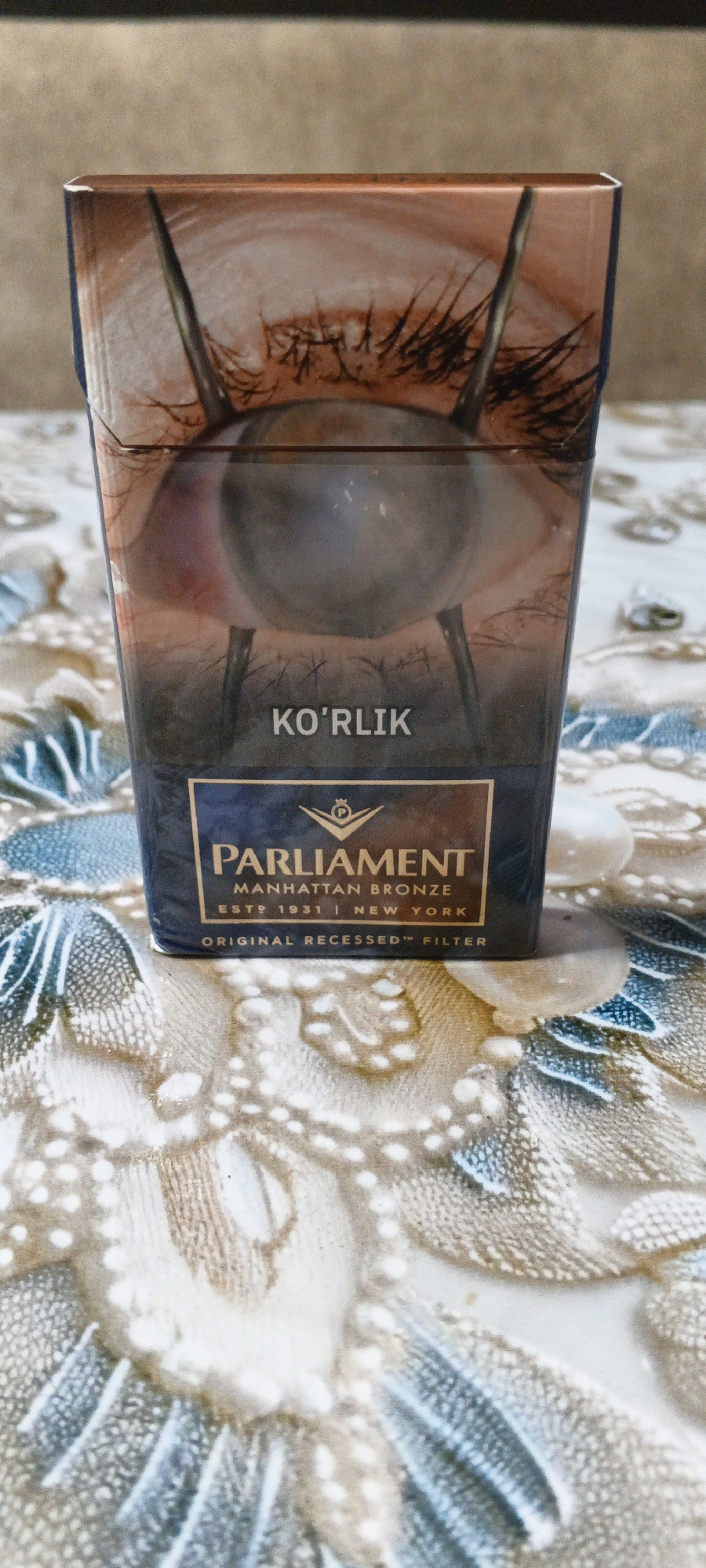 The image displays a close-up, vertical, eye-level view of a pack of Parliament cigarettes. The pack is positioned centrally, resting on a patterned surface that appears to be a tablecloth or placemat with a floral design. The cigarette pack has a dark blue panel at the bottom, featuring the Parliament logo and text that reads "PARLIAMENT MANHATTAN BRONZE ESTD 1931 NEW YORK ORIGINAL RECESSED FILTER". Above this, the pack has an image of a human eye with dark eyelashes and the text "KO'RLIK" in white capital letters. The upper portion of the pack shows more of the eye and surrounding skin tones. The lighting is direct, creating some highlights and shadows on the pack and the patterned surface. The background is blurred and appears to be a neutral, textured wall. There are no people or specific city landmarks visible in the image that would indicate a location within Mustakillik, Uzbekistan.