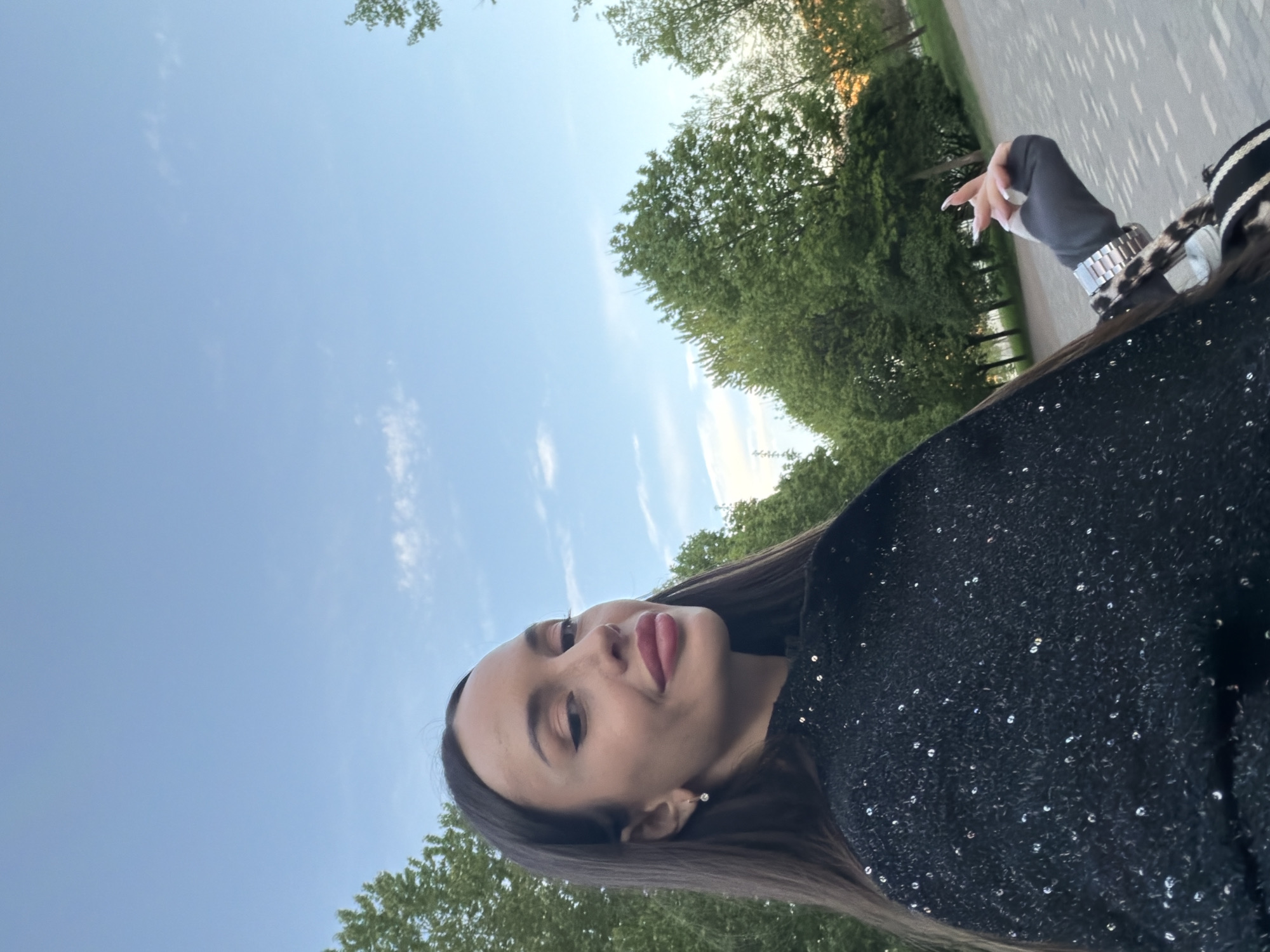 The photograph is an eye-level portrait of a woman in an outdoor setting, likely in Saint Petersburg, Russia, as suggested by the potential park environment. The woman has long brown hair, fair skin, and appears to be wearing a dark sequined top or dress. Her lips are painted a red or pink hue. She is glancing off to the left. She is wearing a watch and a glove. Behind her is a blue sky with scattered clouds, and green foliage, possibly trees in a park. To the right is a stone pathway or pavement.