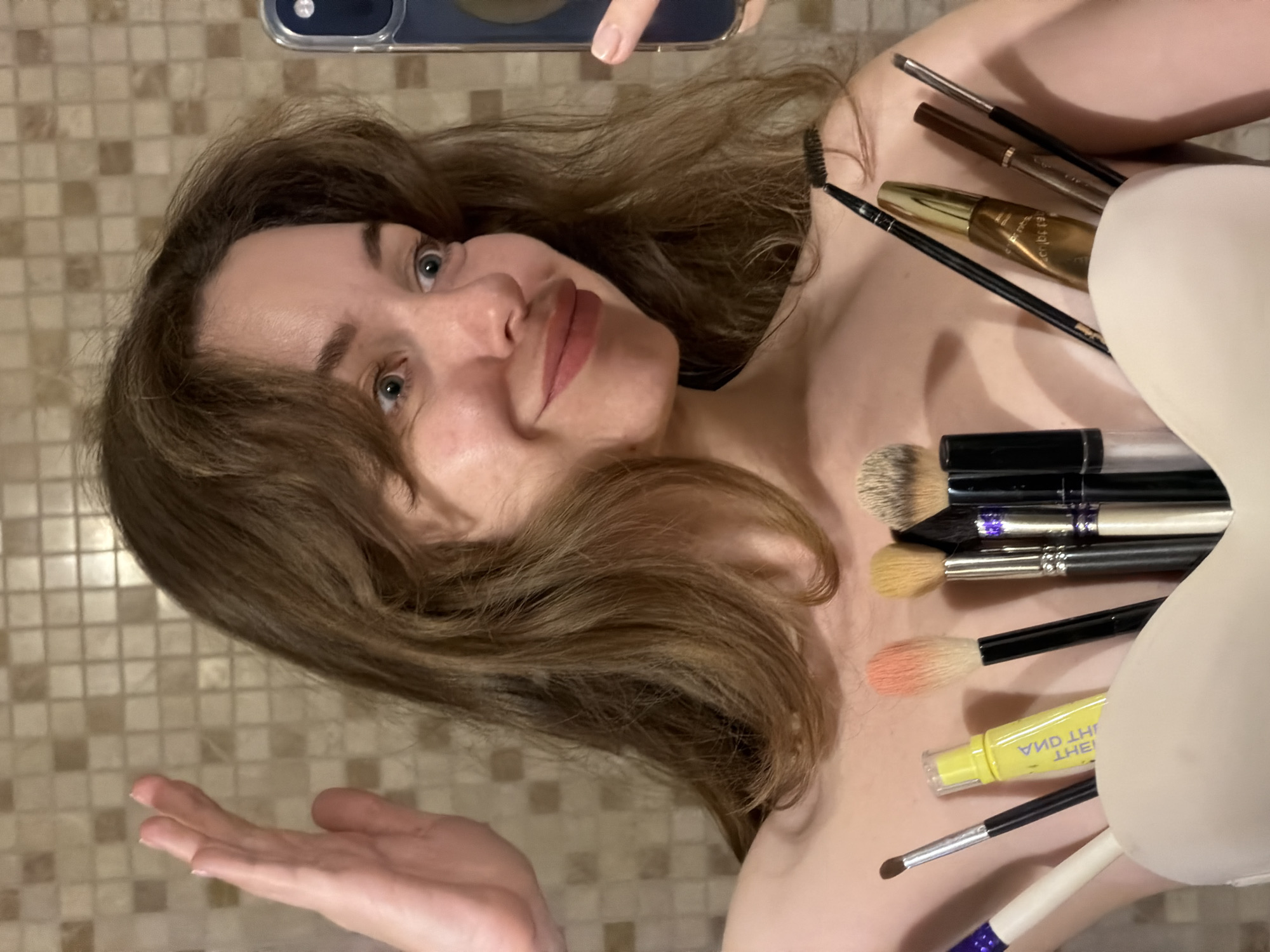 A woman is lying down, possibly in a bathroom given the tiled background. She is holding a phone, presumably taking a selfie. Assorted makeup brushes and a yellow tube of makeup are spread on her chest. The location is not verifiable as Mishutkino, Russia.