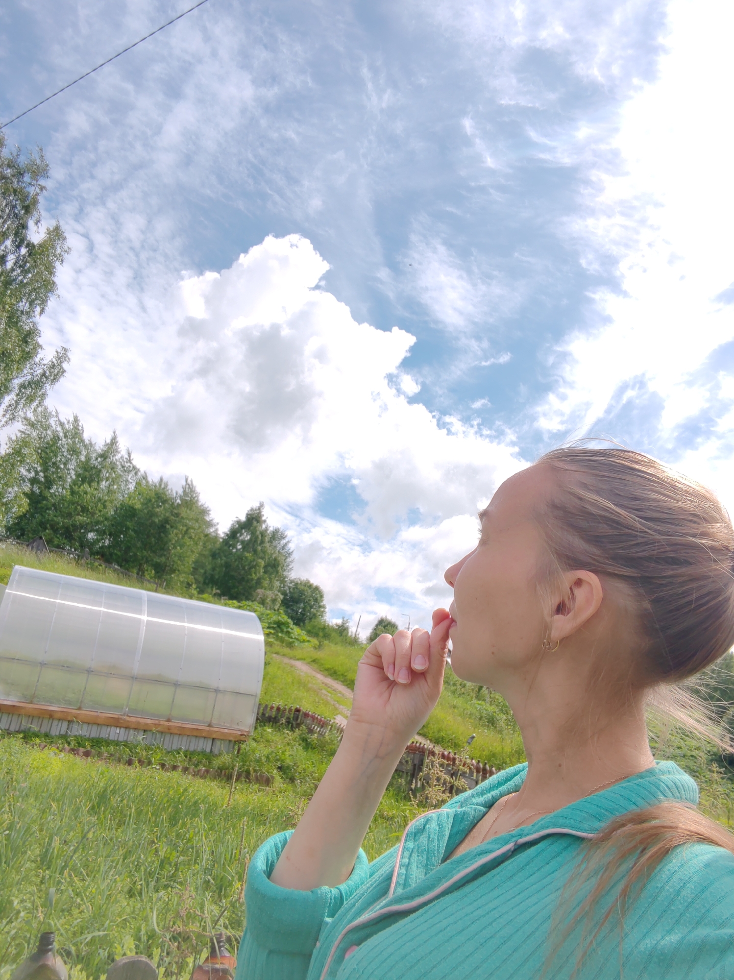 A young woman with blonde hair pulled into a bun is shown in profile, looking upwards and to the left. She is wearing a turquoise robe with pink trim and a delicate gold necklace. Her right hand is near her mouth, with her thumb and forefinger touching. The background features a cloudy blue sky. In the middle ground, there is a greenhouse, lush green vegetation, and trees. The scene appears to be a rural or suburban outdoor setting, possibly a garden or backyard, in the city of Chukhlem, Russia.