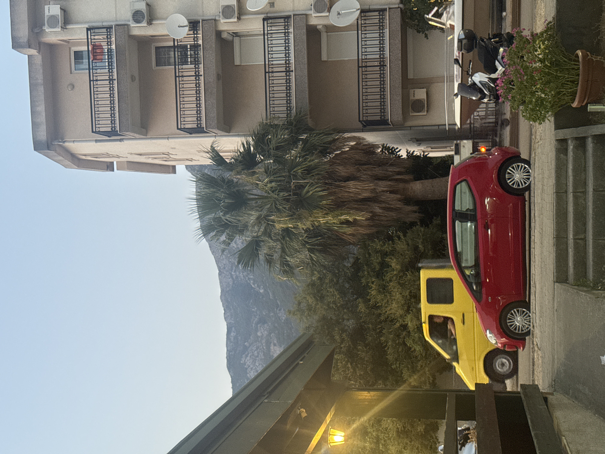 In Sutomore, Montenegro, a street scene unfolds with a multi-story building dominating the right side of the frame, characterized by its beige facade, multiple balconies with black railings, and several air conditioning units. To the left, a structure with a green roof and an illuminated yellow lamp is partially visible. The foreground features a red hatchback parked on the street, its brake light illuminated, and a yellow van with a person inside. Behind the vehicles, a large palm tree with dried fronds stands prominently, obscuring the view of dense green foliage and a hazy mountain range in the background under a clear sky. A white motorcycle with a black seat and a black storage box is parked near the building, adjacent to a potted plant with pink flowers. A boat is also visible in the background, parked near the building.
