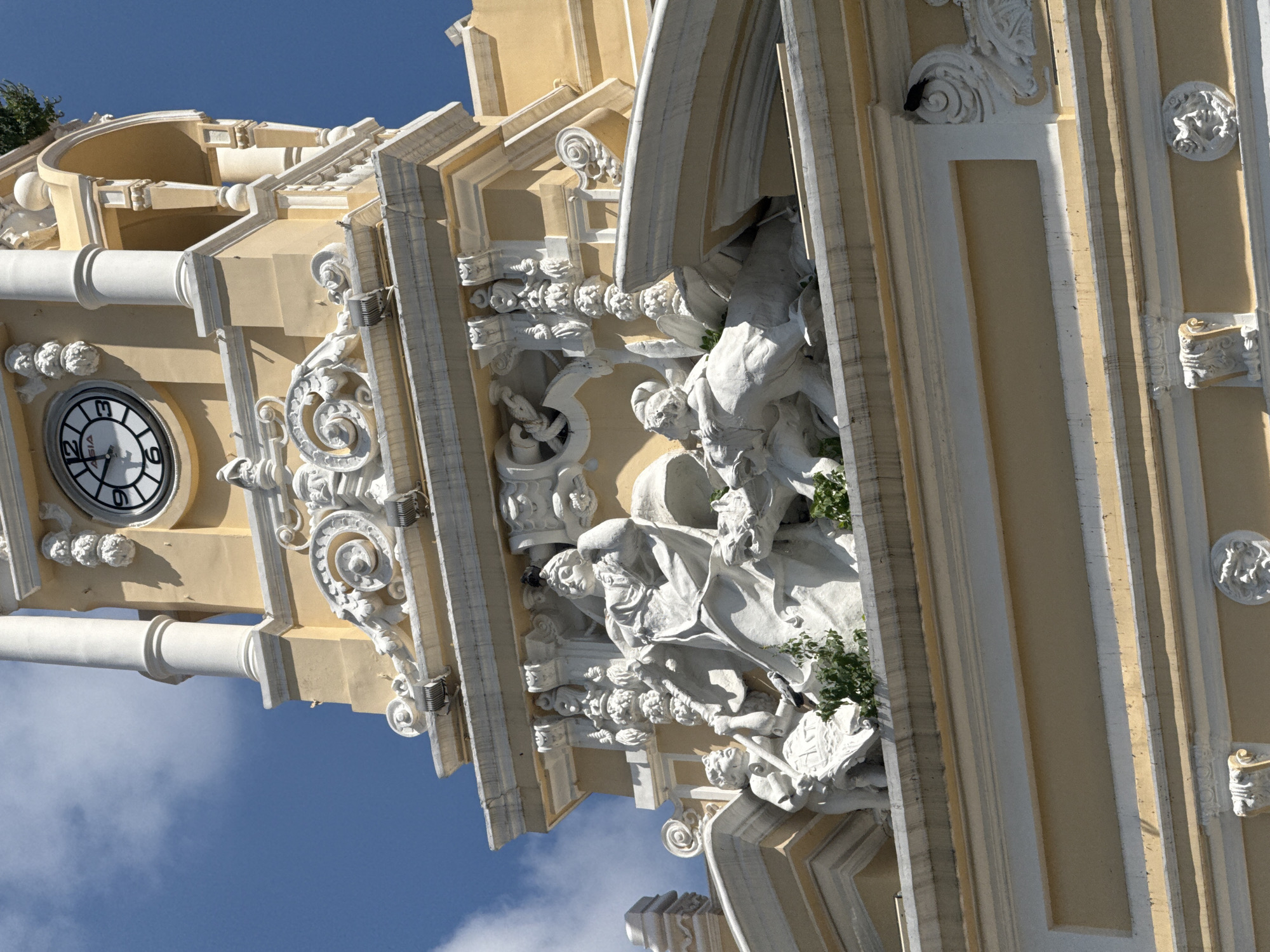 The image shows a close-up view of the facade of a light yellow building with ornate white architectural detailing, set against a bright blue sky with scattered white clouds. Prominently featured is a clock tower with a white-faced clock displaying roman numerals. Below the clock, a detailed white sculptural frieze depicts several figures, including what appears to be a seated woman and two other figures, possibly children or mythological beings. There are also elements of greenery growing on the facade, suggesting the building may be old or in a tropical climate. The building's architecture is grand and classical, with columns, cornices, and decorative carvings. Based on contextual information, this building is likely the Saigon Central Post Office in Thủ Đức, Vietnam, a notable landmark known for its French colonial architectural style and prominent clock tower.