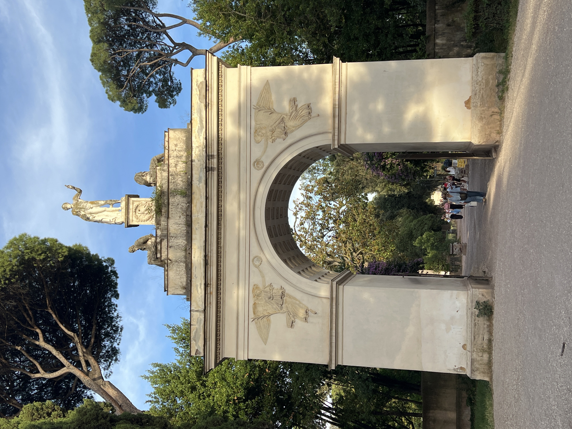 The image displays a grand, weathered stone archway or gate, likely a monumental entrance, situated amidst lush greenery under a bright blue sky. The structure, predominantly light beige or cream-colored, shows signs of age with some darker stains and weathered patches. It features a large central arch opening with a patterned, possibly coffered, ceiling within.
Perched atop the archway's left side are two classical statues: a prominent standing male figure with an outstretched arm, mounted on a tall pedestal, and a smaller, reclining figure positioned below it on the structure's base. Adorning the vertical surfaces flanking the arch opening are intricate bas-relief carvings depicting winged, draped figures. One figure, located on the upper left, appears to hold a circular object or wreath, while another on the lower left holds what looks like bundled fabric or multiple items.
To the left of the archway, a large, mature pine tree with distinctive, spreading branches and a dense green canopy reaches towards the sky, its form silhouetted against the bright background. Other trees and green foliage surround the monument.
On the right side of the archway, a paved pathway or road extends, upon which several people are seen walking. They appear to be casually dressed visitors or tourists, some walking towards or through the arch, others moving away.
The lighting suggests late afternoon or early morning, with warm sunlight illuminating parts of the archway and casting gentle shadows. The sky is clear and blue with minimal cloud cover, contributing to a pleasant, historical, and serene atmosphere. The architectural style and prominent pine trees suggest a location within a park or garden in Rome, Italy. No legible text is visible on the structure.