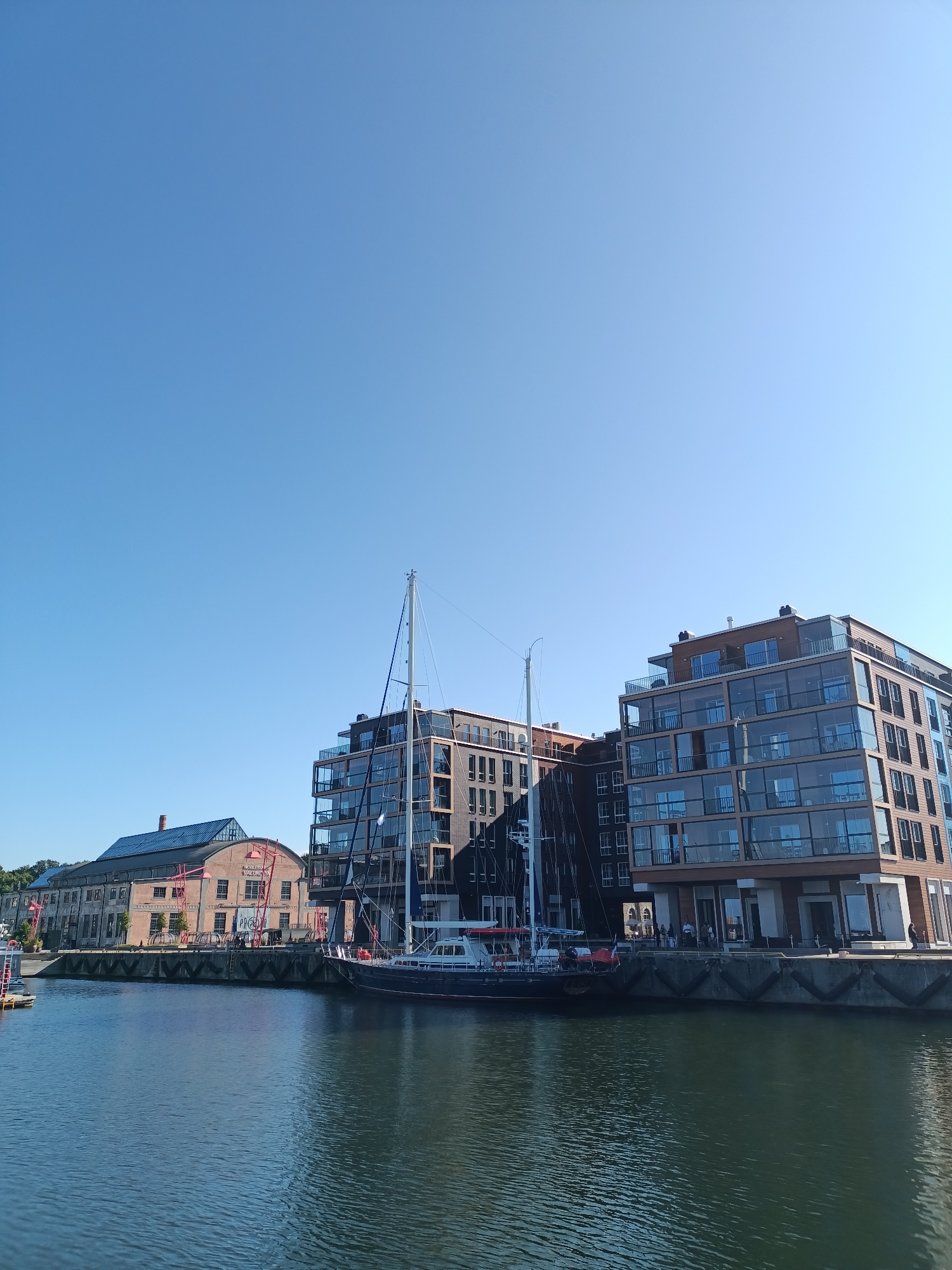 This is a vertical, eye-level, outdoor shot of a harbor in Tallinn, Estonia. In the foreground, the dark blue water of the harbor is visible, with gentle ripples reflecting the clear blue sky above. Along the quay in the midground, there are several modern buildings with dark facades and large glass windows, interspersed with balconies. To the left, there is an older, longer building with a glass roof and red accents. A large sailboat with dark blue hull and white upper works is moored to the quay, with its mast extending high into the sky. Several people are visible on the quay in front of the modern buildings. The scene is bathed in bright sunlight, creating clear shadows.