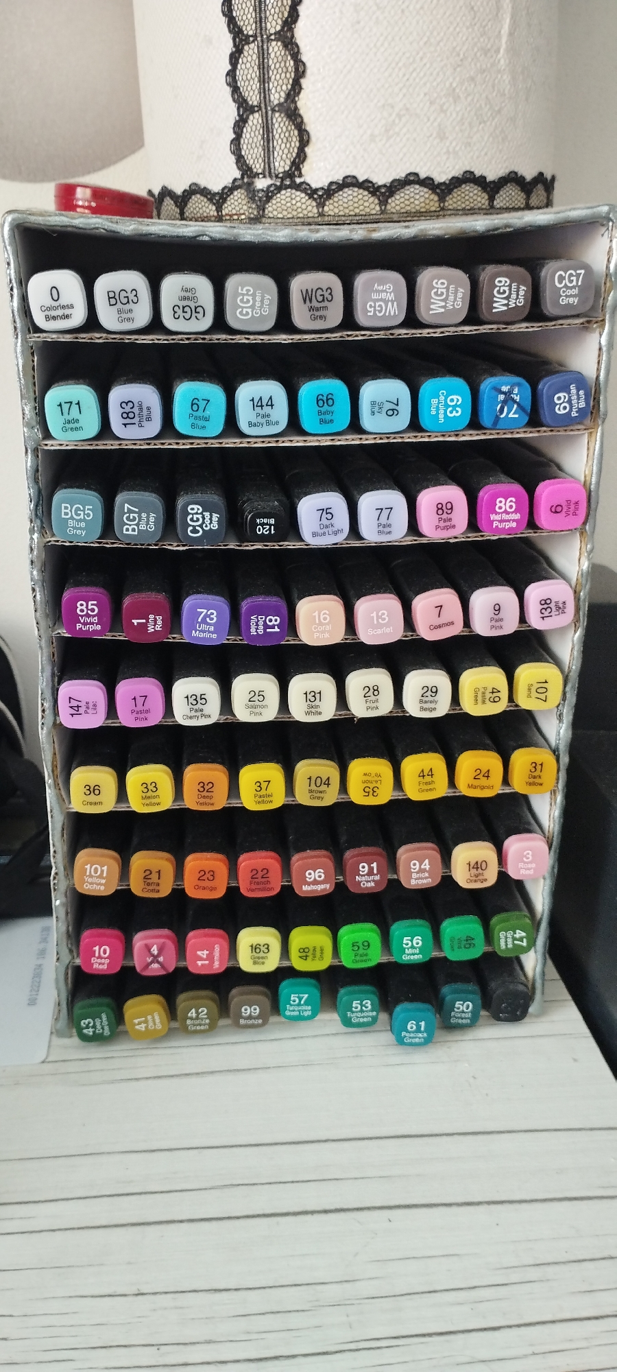 The image shows a rack of markers. The rack is made of cardboard and is divided into several shelves. Each shelf holds a row of markers. The markers are all different colors and have labels with numbers and names. The rack is placed on a wooden surface. There is a white paper roll and a black lace trim in the background. The location in Skidzieĺ, Belarus is not ascertainable from the image.