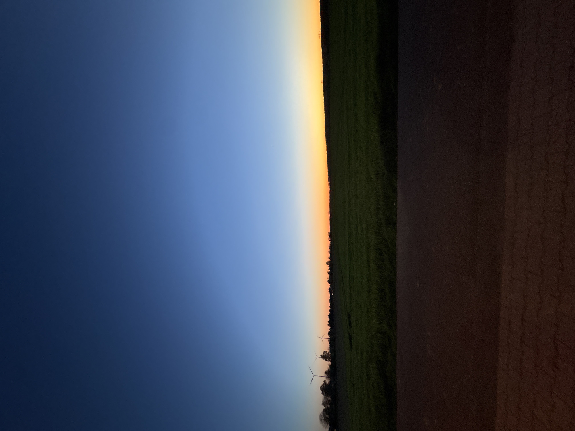 A photograph shows a landscape at dusk. The sky transitions from dark blue to light blue to orange near the horizon. A field of green grass is visible along the horizon line, with a road adjacent to the grass. Three wind turbines are visible on the horizon. The location is in Wegberg, Germany.