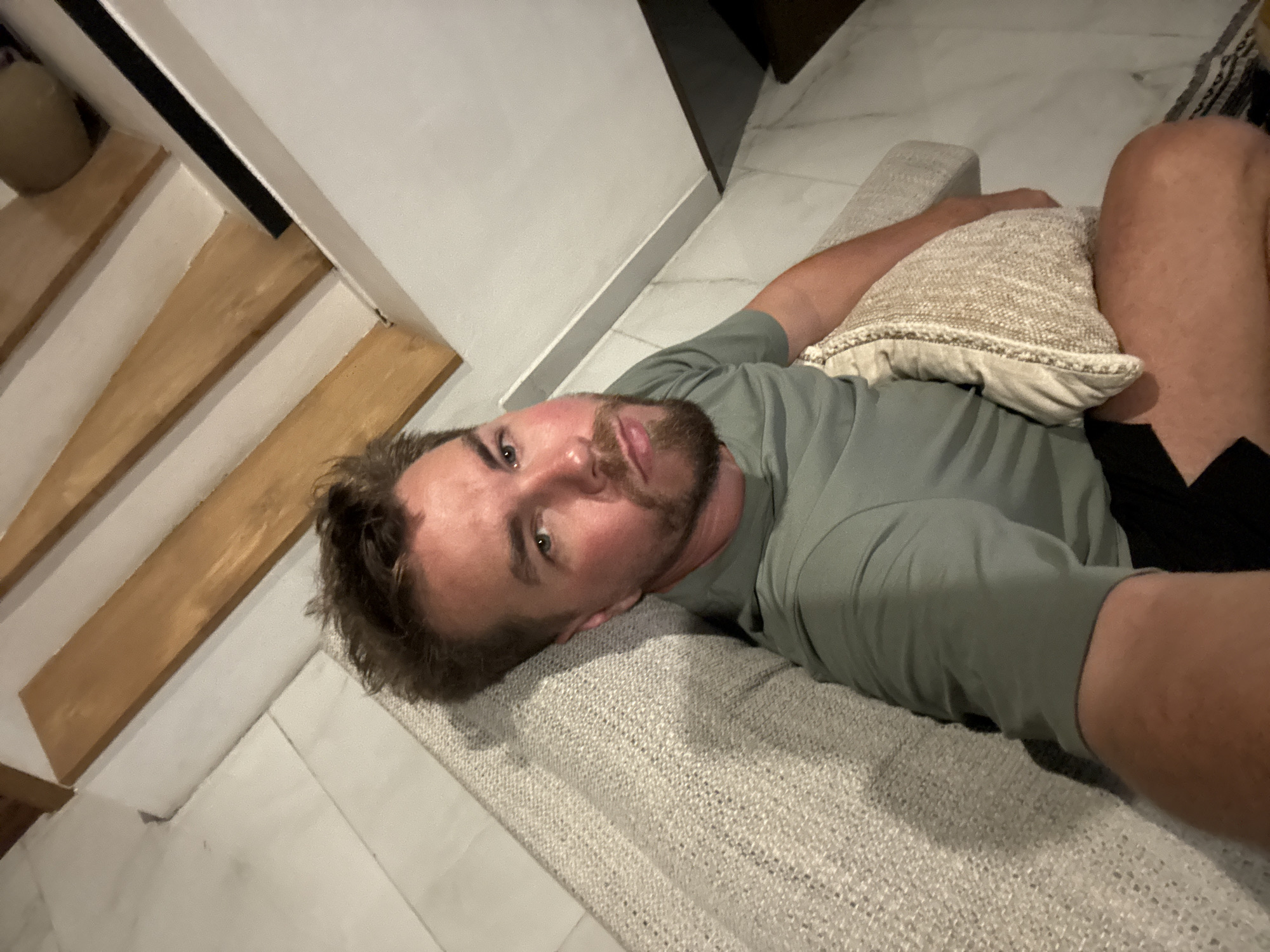A man is reclining on a light-colored, woven sofa with a textured pillow, likely indoors. He is wearing a light green long-sleeved shirt and black shorts. The location is indicated to be North Kuta, Indonesia. To the left of the sofa are stairs with wooden treads and white risers, alongside a white wall. The floor appears to be light-colored tile.