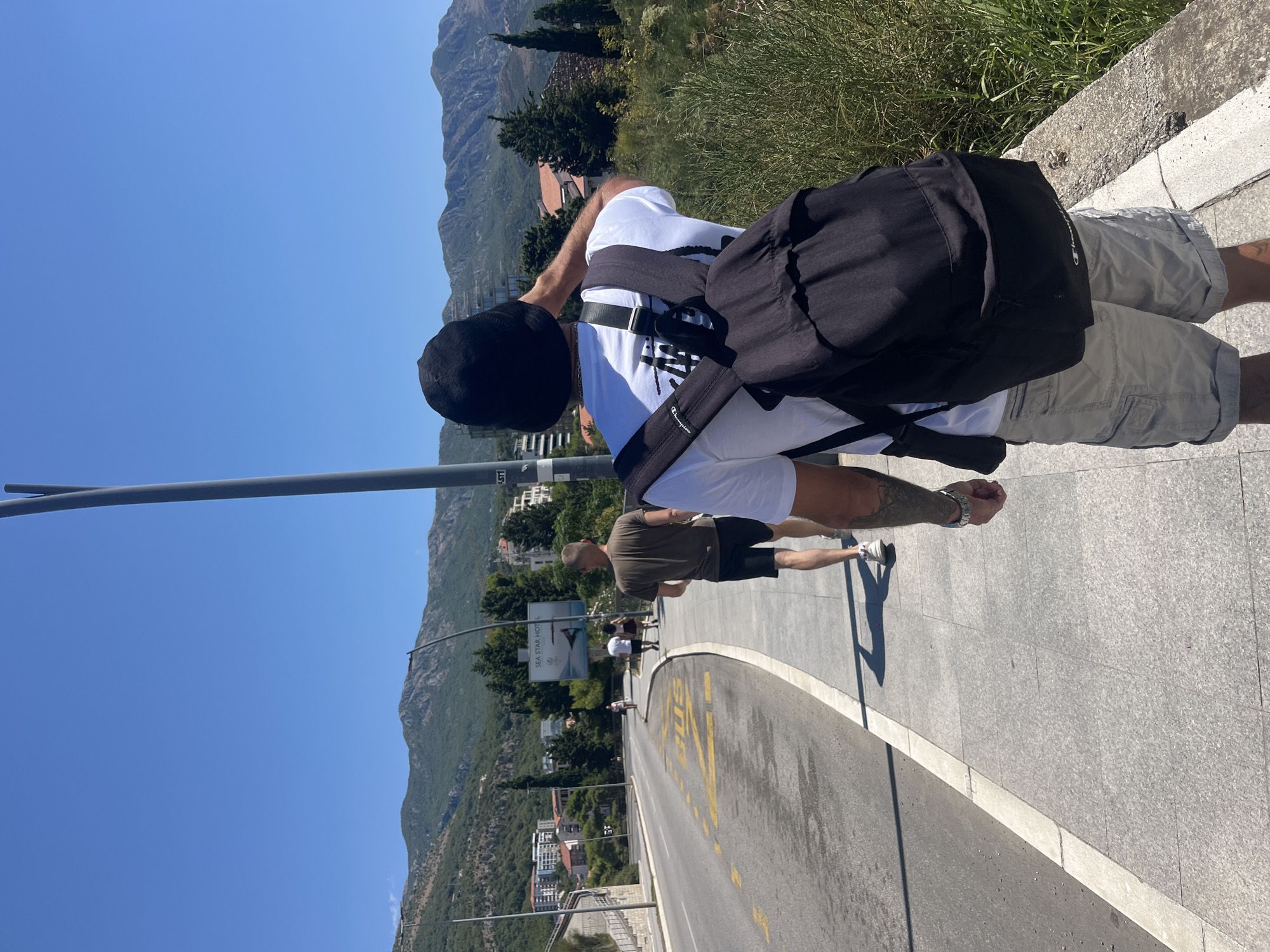 In Boreti, Montenegro, on a sunny day, two individuals walk along a sidewalk adjacent to a two-lane road. The person in the foreground, viewed from behind, wears a white t-shirt with a black graphic, a black beanie, and light-colored cargo shorts. They carry a black backpack and have tattoos on their left arm. To their left, a man in a brown t-shirt and dark shorts walks ahead. The road features yellow "BUS" markings. In the background, a billboard displays "SEA STAR HOTEL." Beyond the road and buildings, a lush, green mountainous landscape stretches under a clear blue sky. The sidewalk is paved with grey tiles, and a concrete barrier separates it from the roadside vegetation.