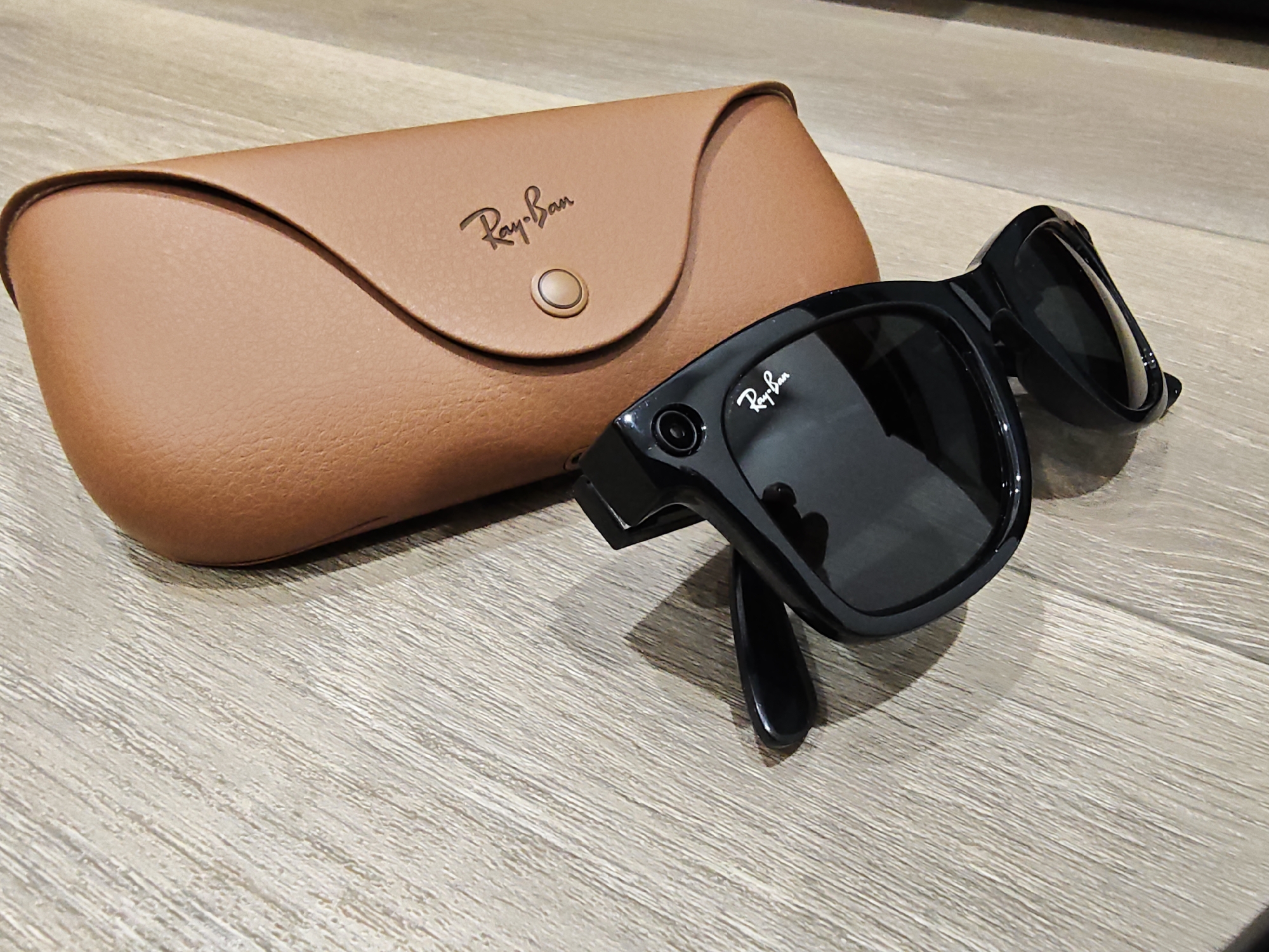 A pair of black Ray-Ban Wayfarer style sunglasses with tinted lenses and a brown leather sunglass case are displayed on a wooden surface. The case is open, revealing a brown interior, and has the Ray-Ban logo embossed on its exterior. The sunglasses are positioned to the right of the case, with one arm folded and the other extended. A subtle reflection of the camera and its surroundings is visible on the lenses. The wooden surface has a light brown, planked texture. This image does not contain any information that would allow for its location to be identified in Calgary, Canada or anywhere else.