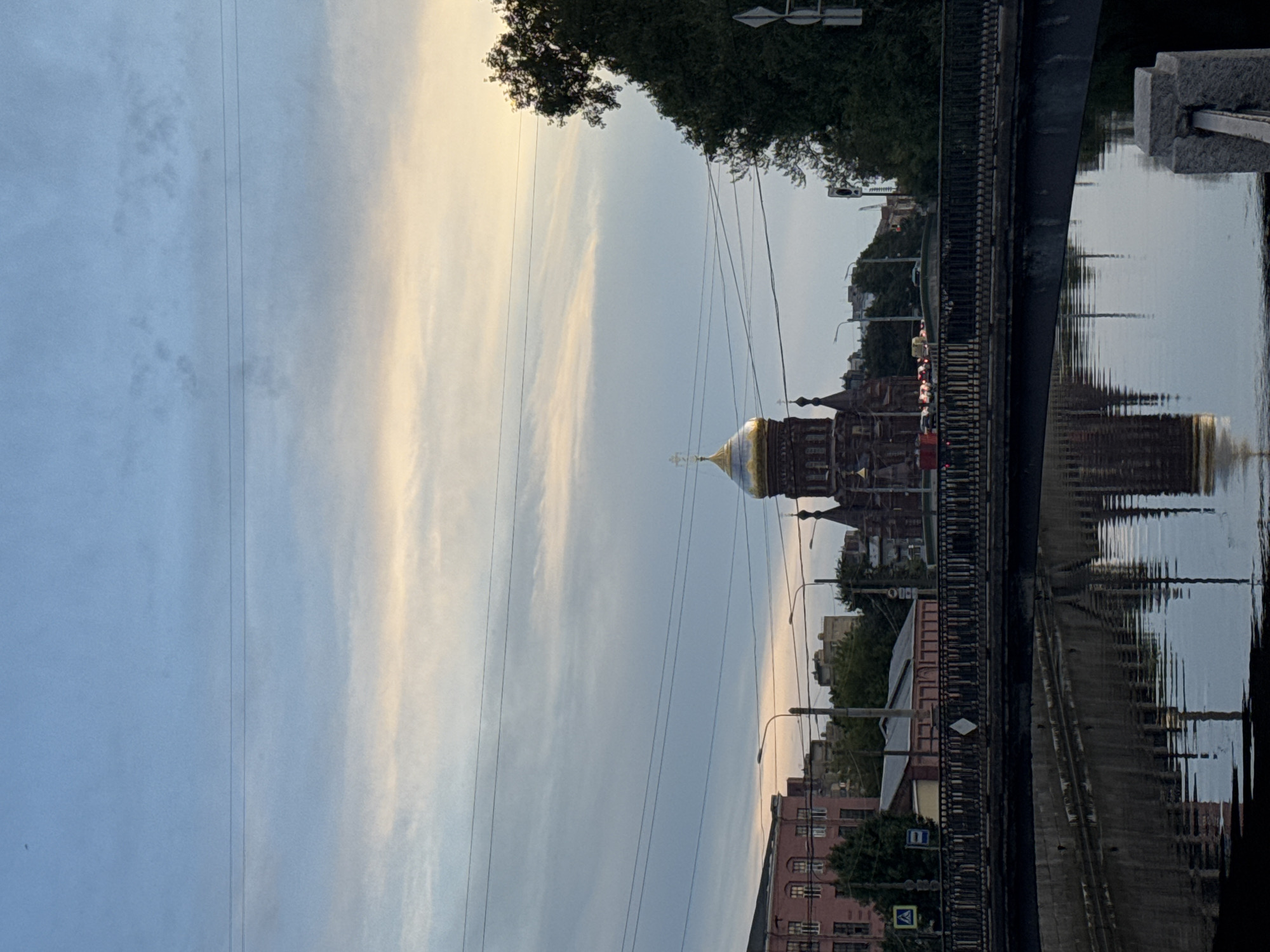 The image depicts a view of a canal with a bridge in the foreground, reflecting the surrounding architecture and sky. A prominent orthodox church with a golden dome stands in the midground, partially obscured by trees and buildings. The sky is a gradient of blue and soft yellow, indicating either sunrise or sunset. The bridge is made of metal with ornate railings, and the canal's water appears calm, mirroring the scene above. Buildings of various architectural styles, including a pink facade on the left, line the canal. Traffic can be seen on the road beyond the bridge, suggesting an urban environment. The overall atmosphere is serene and picturesque. This scene is likely located in Saint Petersburg, Russia, given the architectural style of the church and the presence of canals and bridges common in the city.