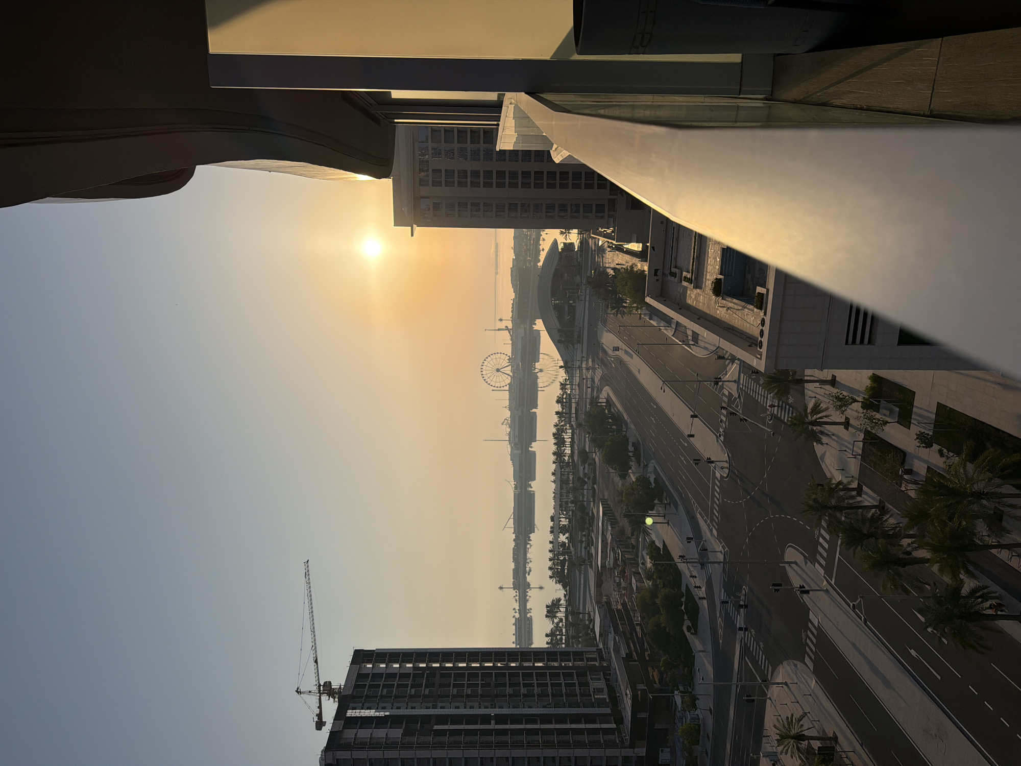 The image shows a sunrise over a city, with a Ferris wheel visible in the distance across a body of water. In the foreground, there is a balcony railing and a building under construction with a crane on top. A street with palm trees lines the waterfront. The sun is low in the sky, casting a warm glow over the scene.