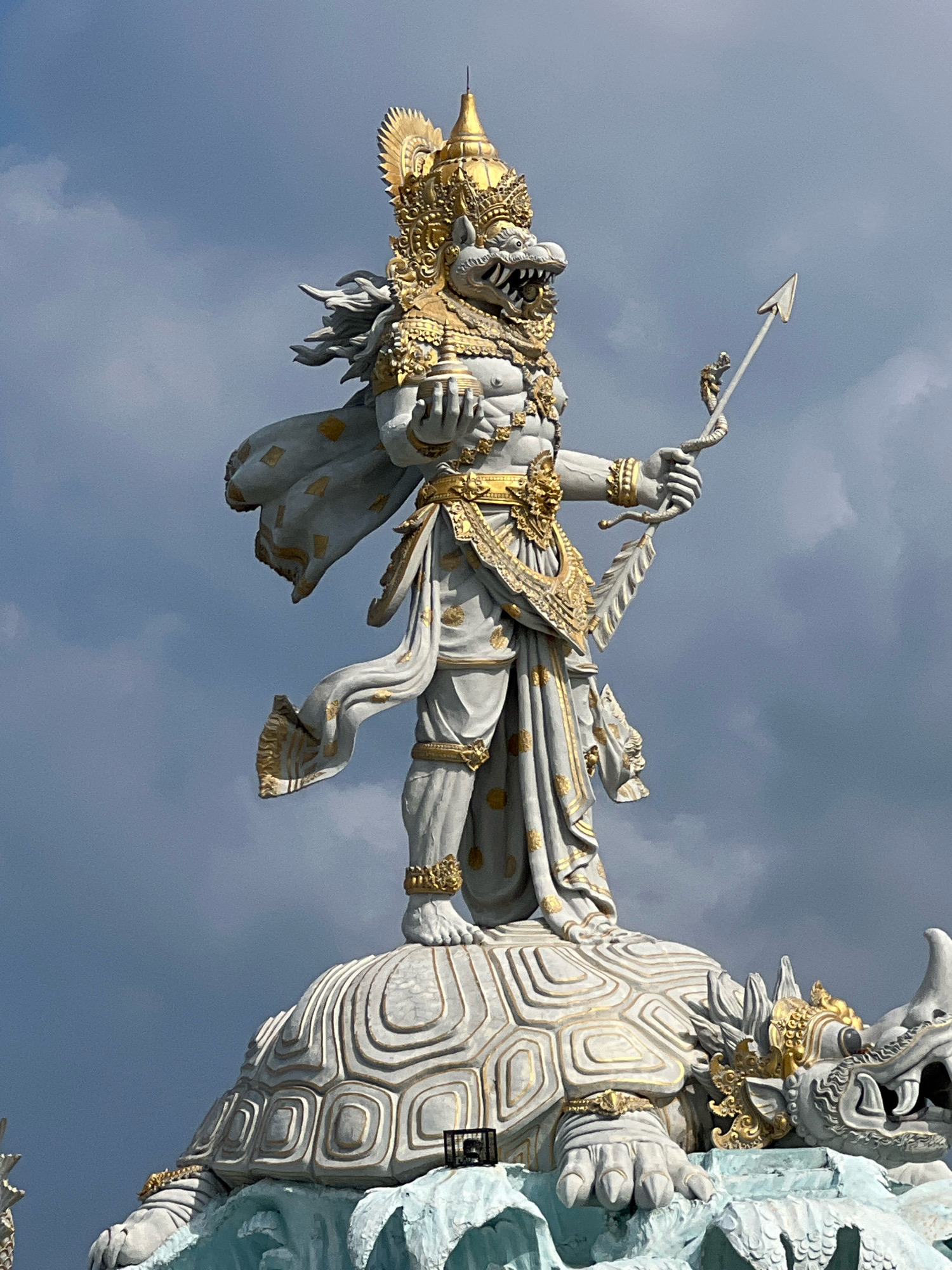 The image shows a large statue of a mythical creature, likely a Hindu deity, standing on a turtle and a dragon. The statue is predominantly white with gold accents, featuring intricate details on its armor, headdress, and clothing. The creature has a fierce expression with sharp teeth and a long, flowing mane. It holds a spear in its right hand and a circular object in its left. The turtle and dragon beneath it are also intricately carved, with scales and sharp claws visible. The statue is set against a dramatic sky with dark, cloudy formations. This statue is located at the Garuda Wisnu Kencana Cultural Park in Kuta, Bali, Indonesia.