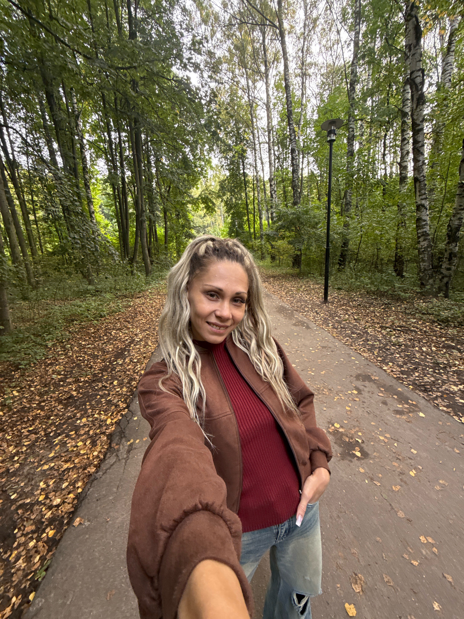 A blonde woman with her hair pulled into two ponytails, wearing a brown jacket over a dark red ribbed top and ripped blue jeans, is taking a selfie while standing on a paved path in a forest. The path is covered in fallen brown leaves, and trees with green and yellow foliage line both sides of the path. A black lamppost is visible on the right side of the path. The location is identified as Rusanovo, Russia.