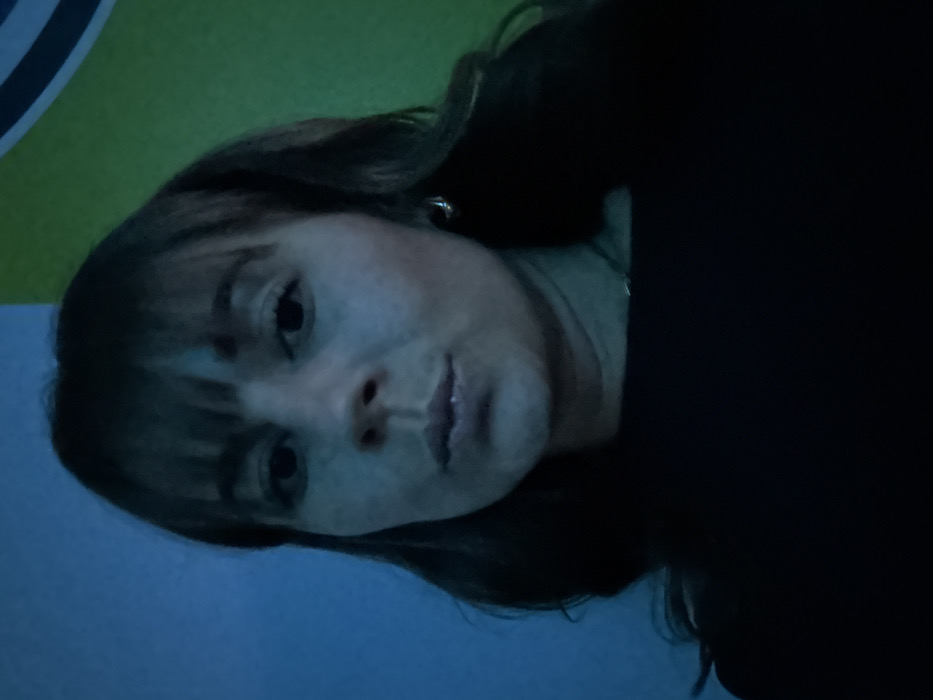 A woman with bangs and dark hair faces the camera. She wears a dark garment, possibly a sweater or shirt. Her skin appears pale, likely due to lighting conditions. She has small, subtle earrings. The background features a mix of blue and green hues, suggesting a wall or other surface with varying colors or lighting. The image is slightly tilted, giving a casual, slightly off-kilter impression.