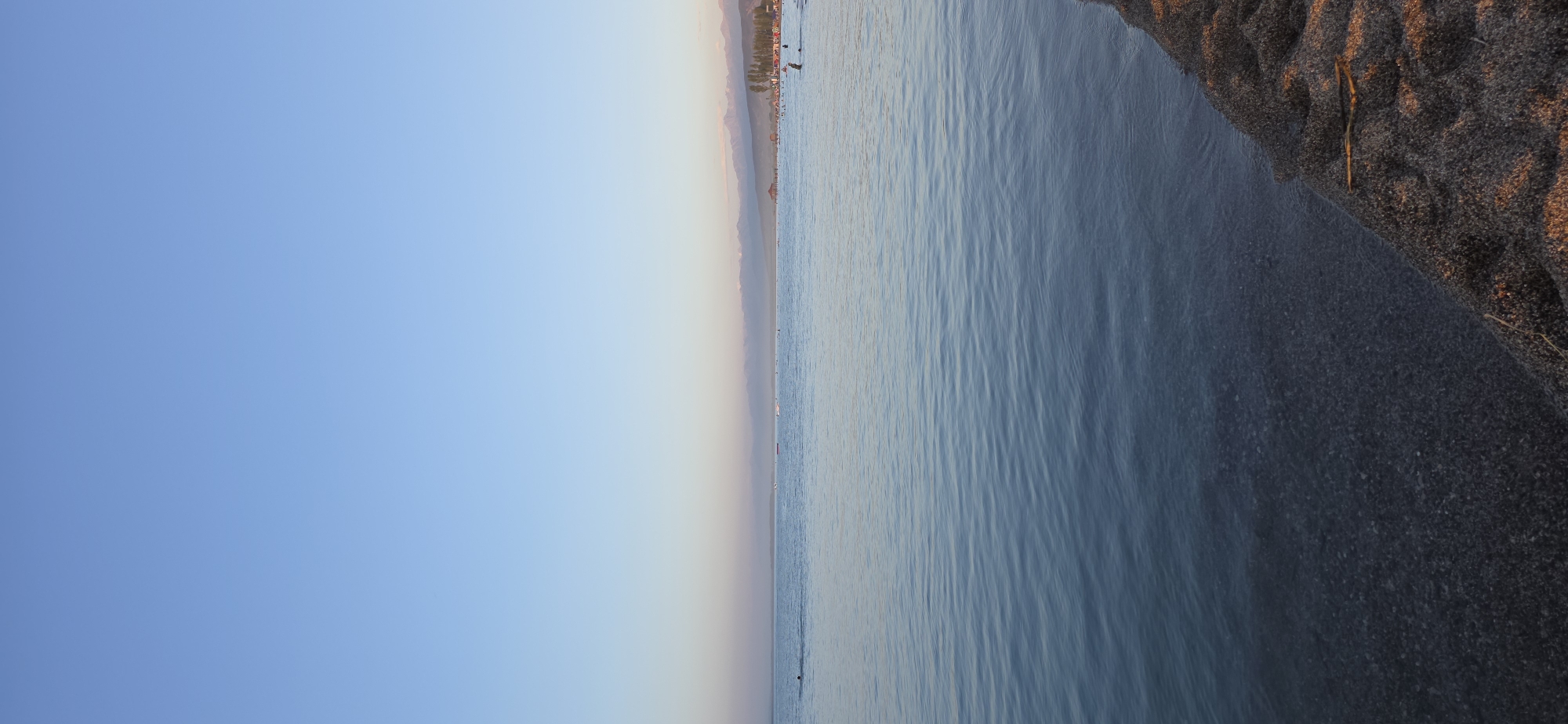 The image depicts a serene beach scene at the city of Boz Beshik, Kyrgyzstan. The foreground features a sandy shore with a few small sticks scattered on it. The water is calm with gentle ripples, and the pebbles beneath the surface are visible. In the distance, a thin line of trees and structures can be seen along the coastline, with a mountain range forming the backdrop under a clear, pale blue sky. Several people are present in the water, some swimming and others appearing to be relaxing, while a few more are visible on the distant shore. The overall atmosphere is peaceful, suggesting a tranquil day at the beach.