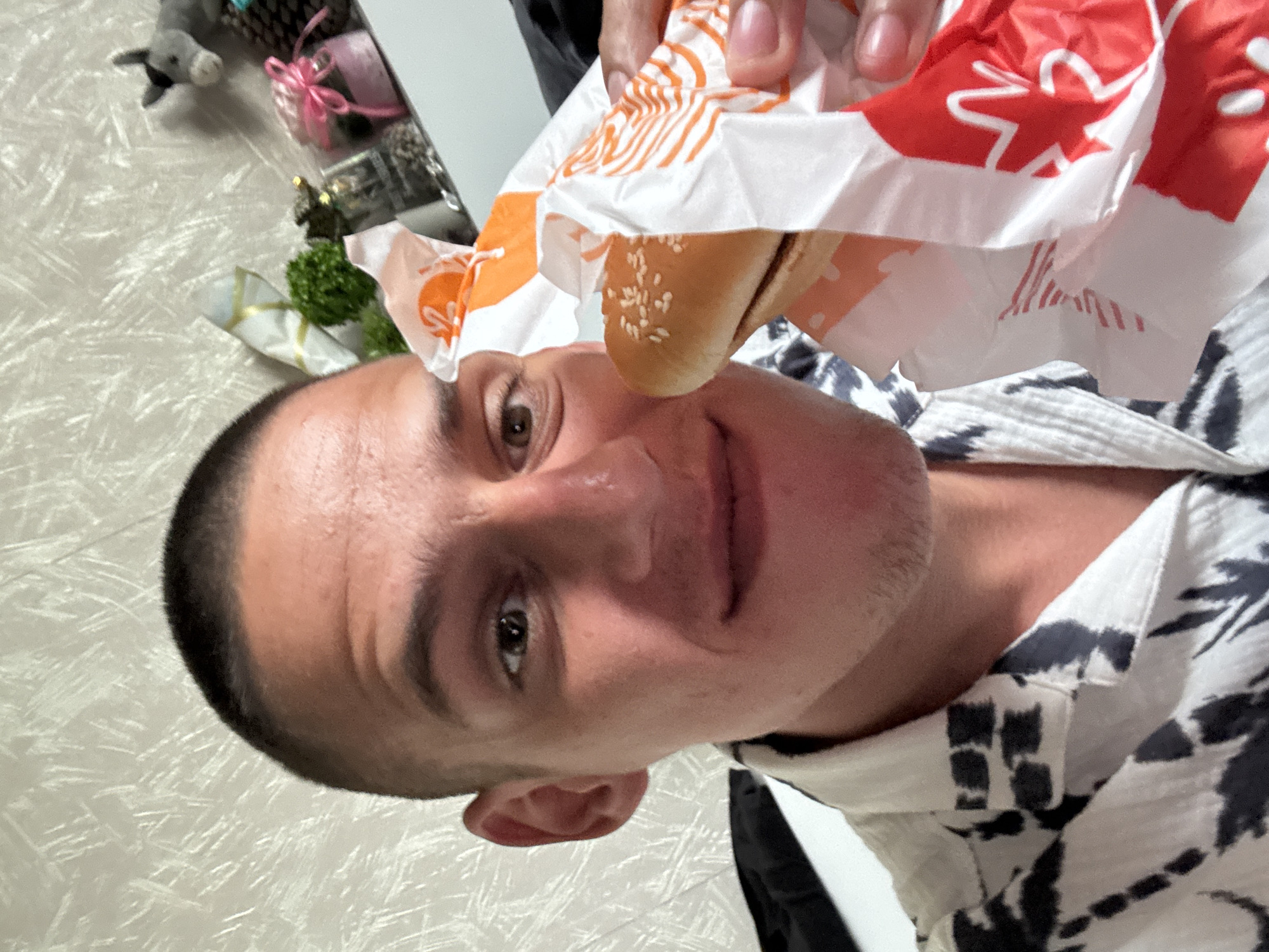 A man holds a burger in front of his face, with the top bun touching his nose. The burger is in a Burger King wrapper. The man has a shaved head and is wearing a black and white shirt. The background is a wall. The location cannot be determined with just the image provided.