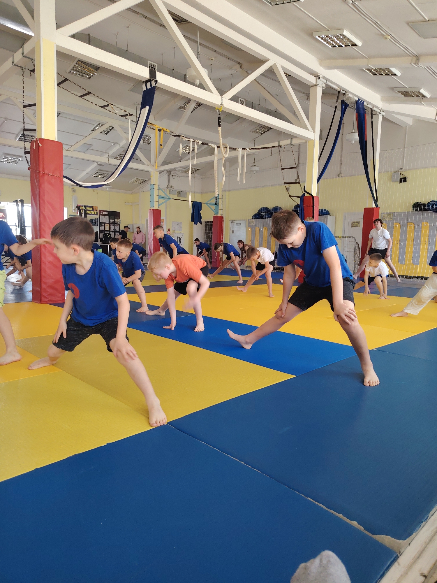 The image shows a group of young people stretching in a gym in Chaikovsky, Russia. The gym has a high ceiling with exposed support beams, and the floor is covered with blue and yellow mats. The children are mostly dressed in blue T-shirts and black shorts, and are barefoot. They are performing a stretching exercise, with one leg extended and the other bent. An adult male is also standing near the wall, possibly instructing the children. Ropes and gymnastic equipment are visible in the background.