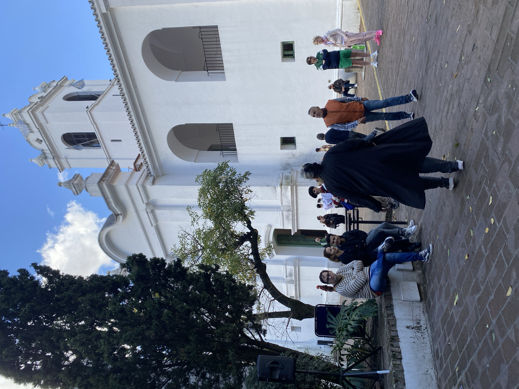 The image depicts a scene in Buenos Aires, Argentina. In the background is a white colonial-style building, likely a historical structure, possibly associated with a church, given its architectural features such as the tower. A crowd of people is gathered around the building. Some are sitting on a low wall or ledge in the foreground. Others are walking around or standing further back. A large tree is in front of the building, partially obscuring the view. There is also a speaker on a stand. The image seems to be showing an outdoor event or gathering, possibly a tour. The sky is partly cloudy. The ground is paved with bricks or tiles.