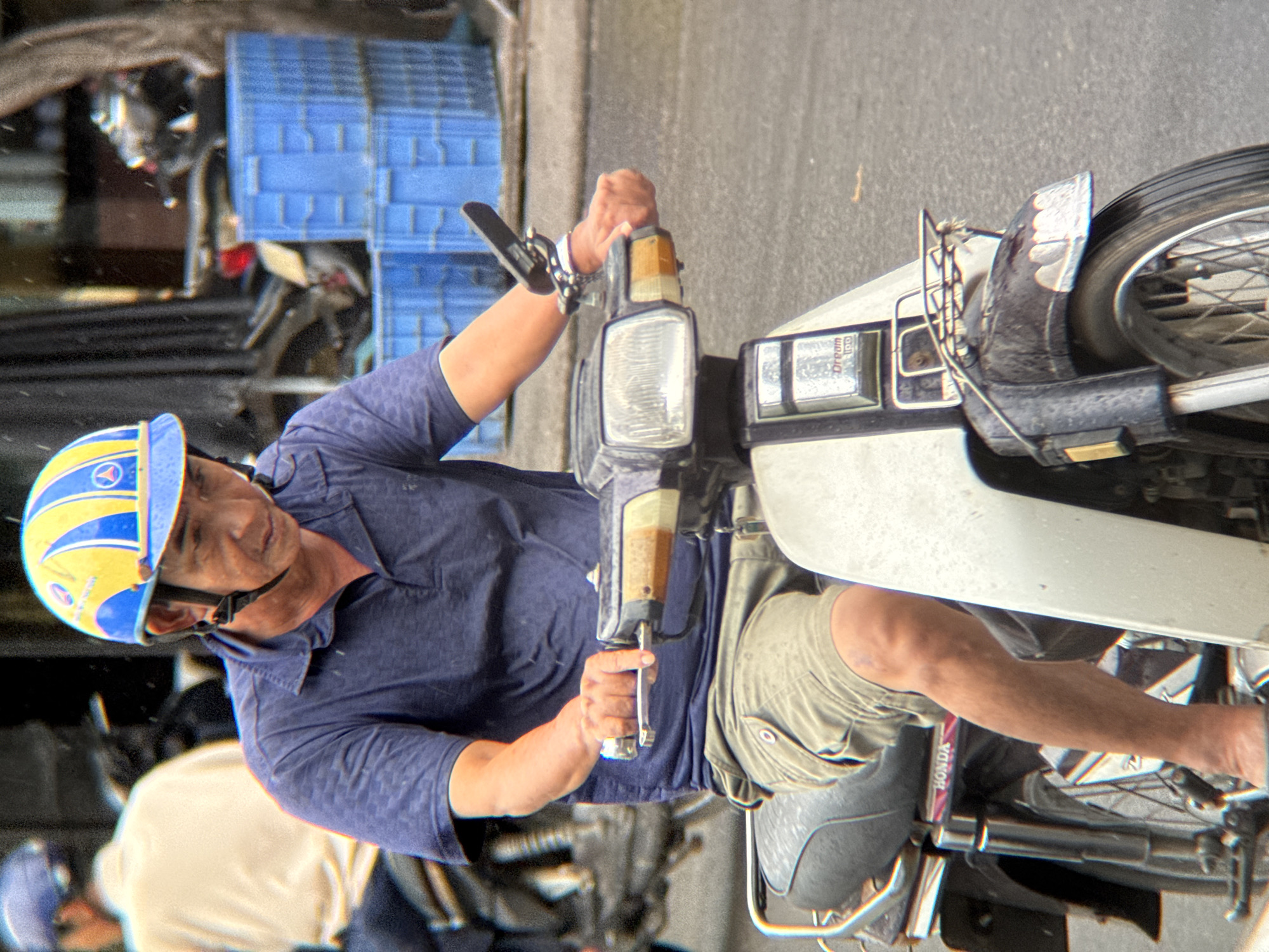 A man in a blue polo shirt and khaki shorts rides a scooter on a paved street. He is wearing a yellow and blue helmet. The scooter has a white front panel and a black fender. In the background, there are blue crates stacked on top of each other and a blurry figure on another scooter. The scene appears to be in a city, possibly Nha Trang, Vietnam, given the style of the scooter and the presence of blue crates commonly used for transport in Southeast Asia.