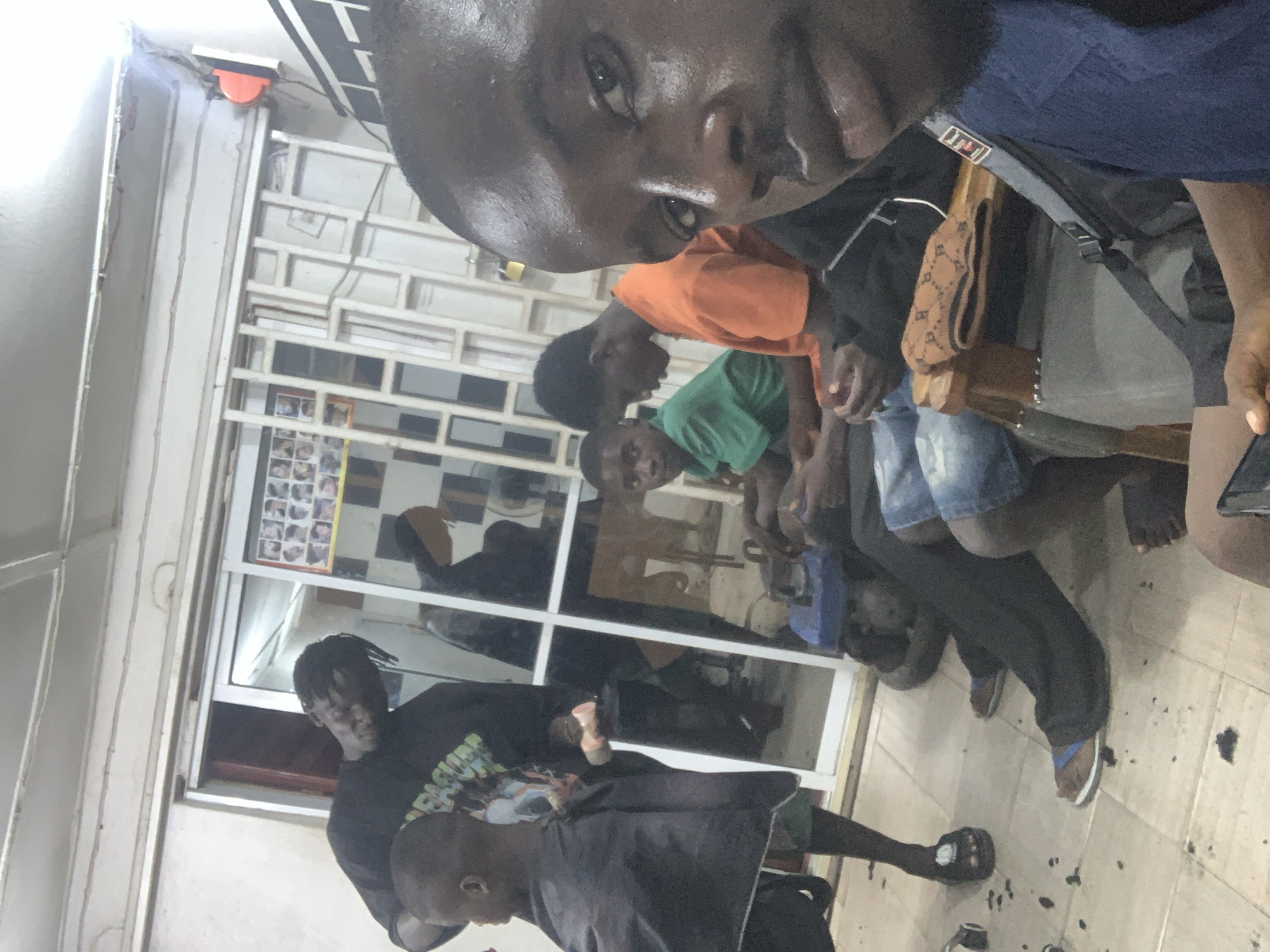 A dimly lit interior shot shows a barber and his customers. The location is indicated as being in Jos, Nigeria. At the left of the shot is a barber wearing a black t-shirt with dreadlocks. He is focused on cutting the hair of a young boy wearing a black barber cape. Several customers are sitting in the chairs at the back watching. One boy is wearing an orange t-shirt, and one a green t-shirt. The foreground is dominated by a dark-skinned man with a beard, presumably holding the camera. The floor appears to be tiled with a light-colored material, covered with hair clippings.