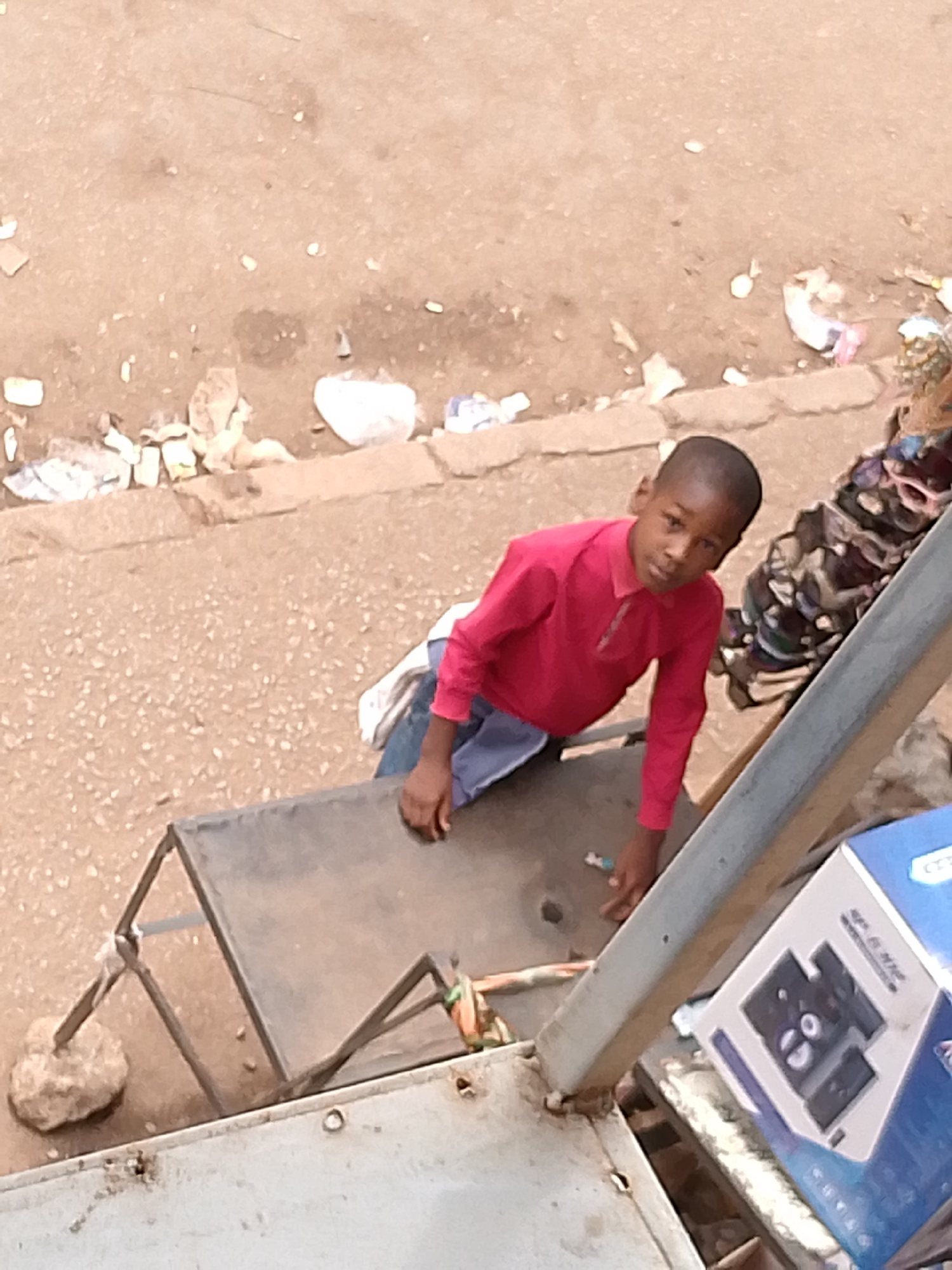A young person in a red shirt and blue pants is standing on top of a metal structure. The structure is propped up by a rock. Next to the metal structure is a blue box. The scene appears to be set in an urban environment. There is garbage on the ground next to a sidewalk.