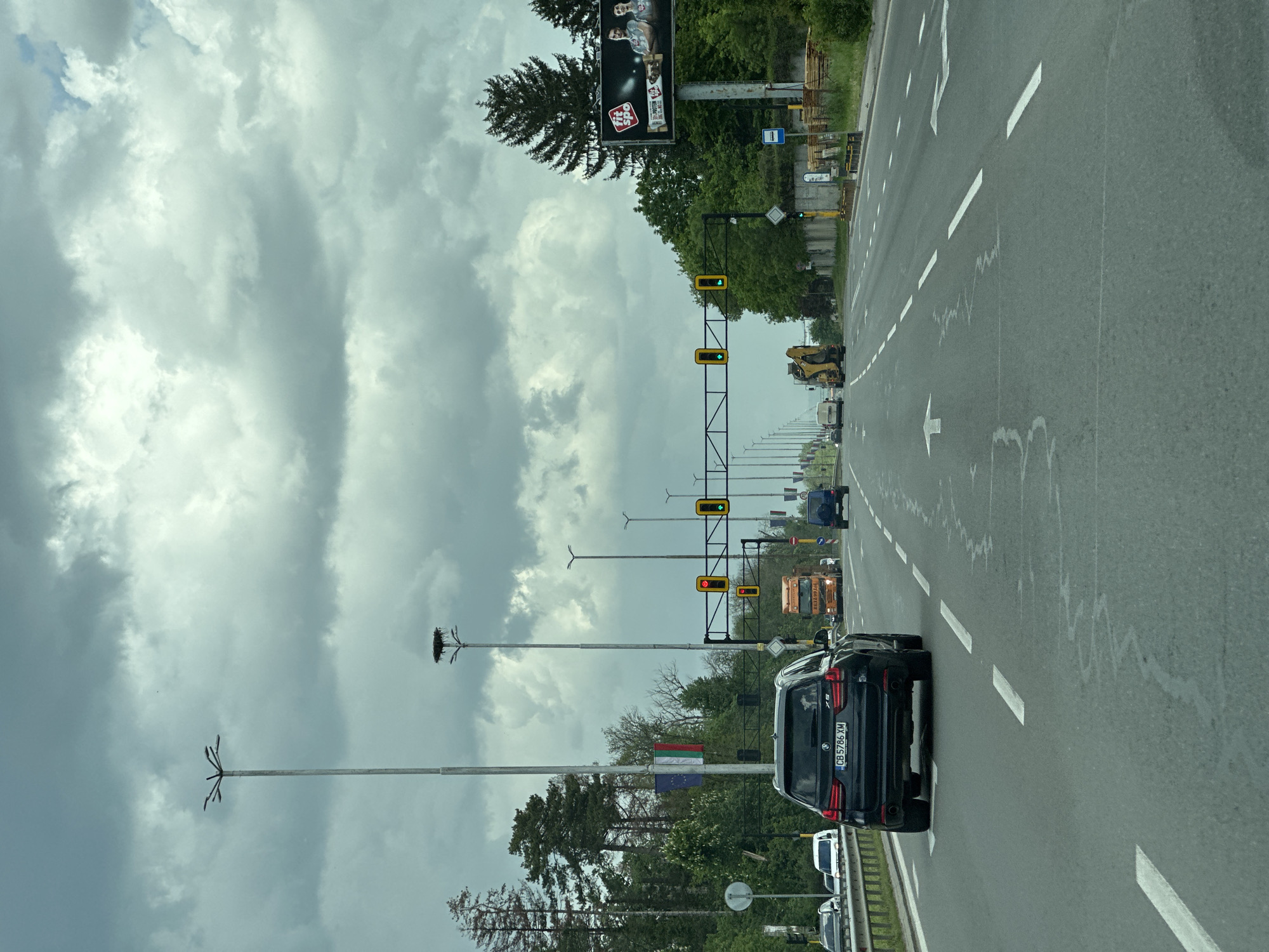 The image depicts a road in what appears to be the city of Sofia, Bulgaria. The photograph is taken from a vehicle traveling along the road. In the foreground, a black SUV is visible with the license plate "CB5786 XM". The road has multiple lanes and lane markings, with an arrow indicating direction.
The road is lined with lampposts, some of which have stork nests on top. There are also traffic lights visible along the road. Further in the background, there is construction equipment and other vehicles.
The sky is overcast with heavy clouds. A billboard is visible on the side of the road. The overall scene is that of a busy road on a cloudy day.