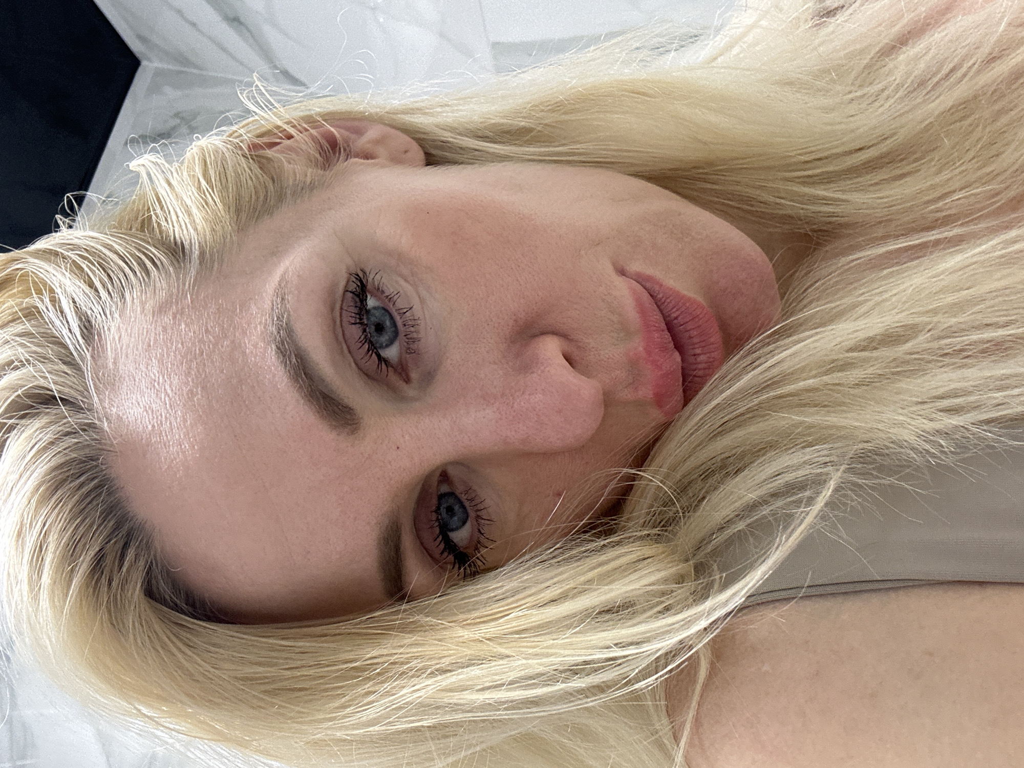 A close-up, slightly angled selfie of a blonde woman looking at the camera. She is wearing a neutral-toned top and has her blonde hair framing her face. Her makeup includes mascara and pink lipstick. The background appears to be a bathroom with white marbled tiles and a black ceiling.