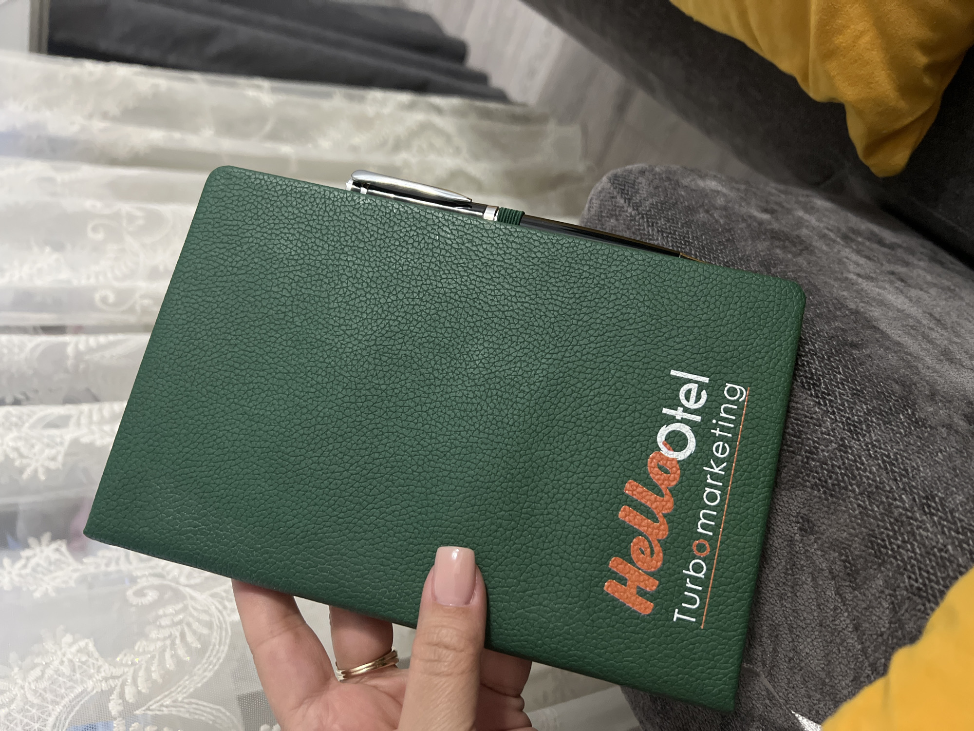 A person's hand, with a light pink manicure and two gold rings, holds a dark green notebook. The notebook has a textured cover and a silver pen clipped to its top edge. The notebook is open, revealing the front cover with text printed in orange and white. The text reads "HelloOtell" in large, bold letters, with "Otell" being in white. Below this, smaller text in orange reads "Turbomarketing". The notebook is resting on a textured, dark gray fabric, possibly a sofa or cushion. In the background, a blurry yellow object, likely another cushion, is visible, along with a white, lacy textile. The scene appears to be indoors, in a casual setting. There are no obvious cues for time of day or weather, but the overall impression is one of a personal moment, perhaps during a break or a moment of reflection, given the relaxed posture and indoor setting. The location context of Kant, Kyrgyzstan, does not provide specific visual cues within the image itself.