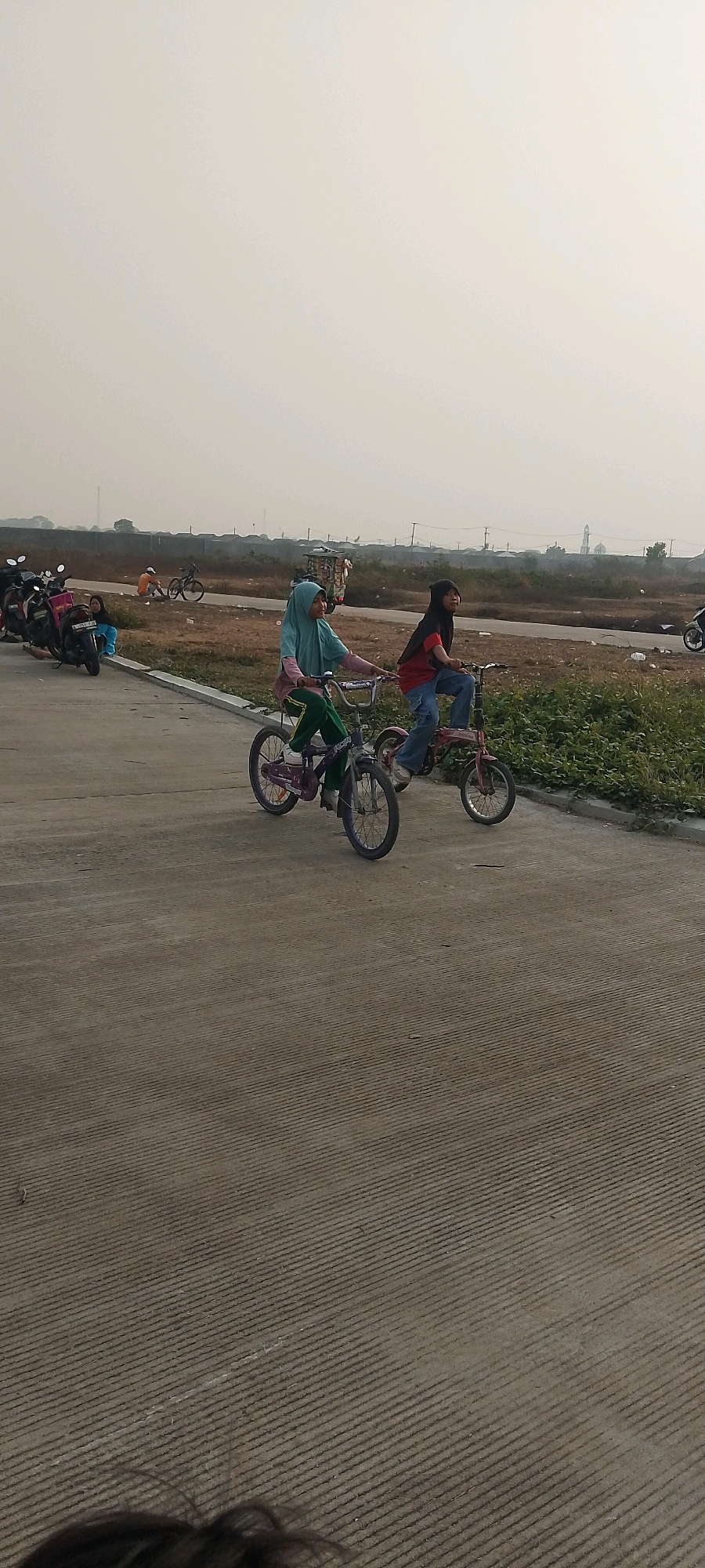 Two young females are riding bicycles on a paved road in Satriamekar, Indonesia. The female on the left is wearing a light blue hijab and green pants. The female on the right is wearing a black hijab and a red shirt with blue jeans. Both bicycles are small, likely for children. In the background, several motorcycles are parked in a line. The environment appears to be an open, undeveloped area with dry grass and some vegetation. There are scattered structures in the distance, possibly indicating a developing residential or industrial zone. The sky is overcast and hazy. The road surface is grey concrete with a textured pattern of parallel lines.