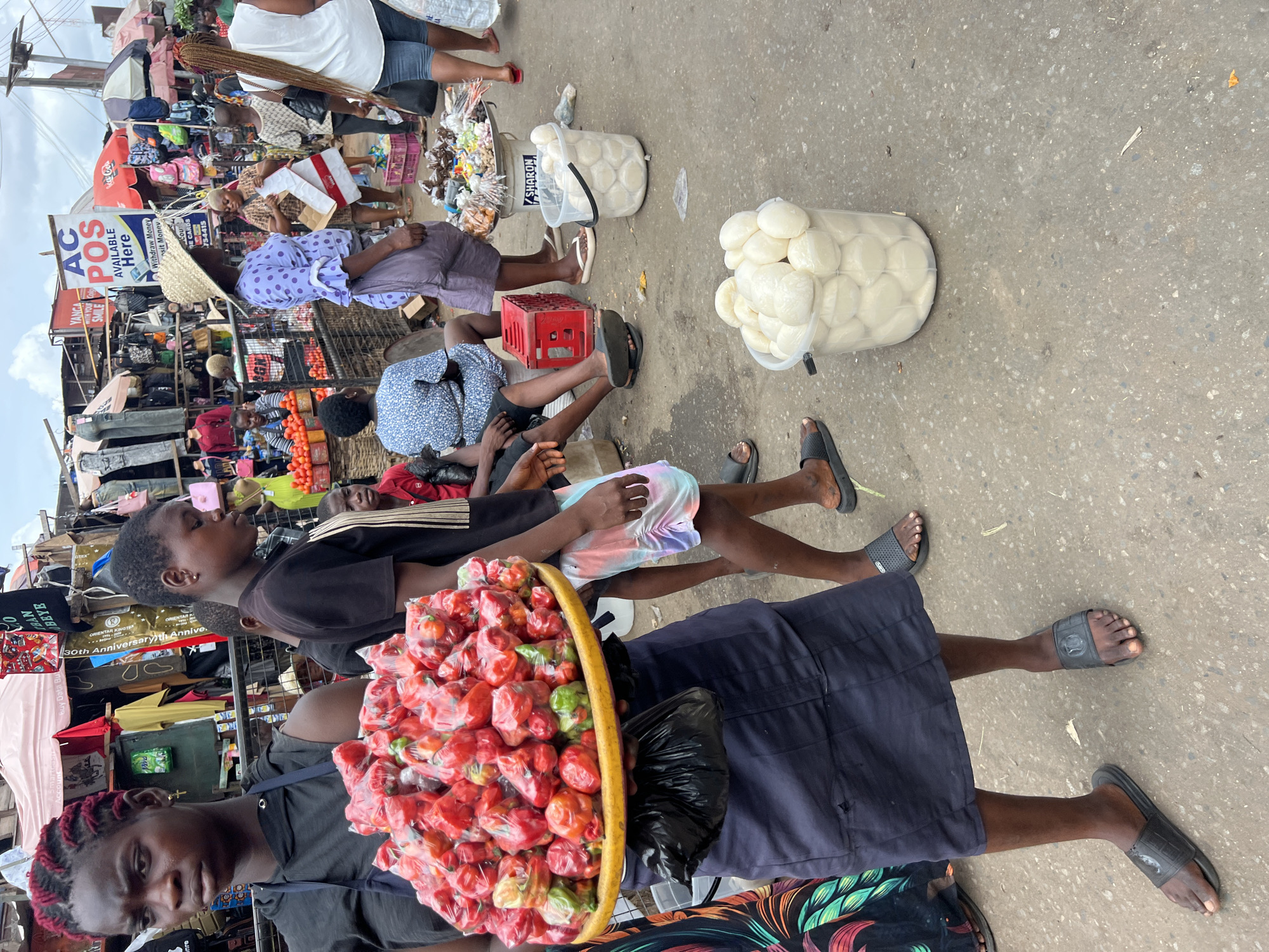 This photograph depicts a bustling street scene in Nkpor, Nigeria, showcasing street vendors and passersby. Two women are prominent in the foreground. One is carrying a basket overflowing with red peppers. Adjacent to the peppers is a black plastic bag. Behind this vendor, a second woman with reddish braided hair is visible. In the mid-ground, a young individual is carrying a container filled with white, round food items. Several other individuals are resting on the ground or seated on crates nearby. The backdrop includes a variety of market stalls selling items such as bags and clothes, with an “AC POS AVAILABLE HERE” sign visible.