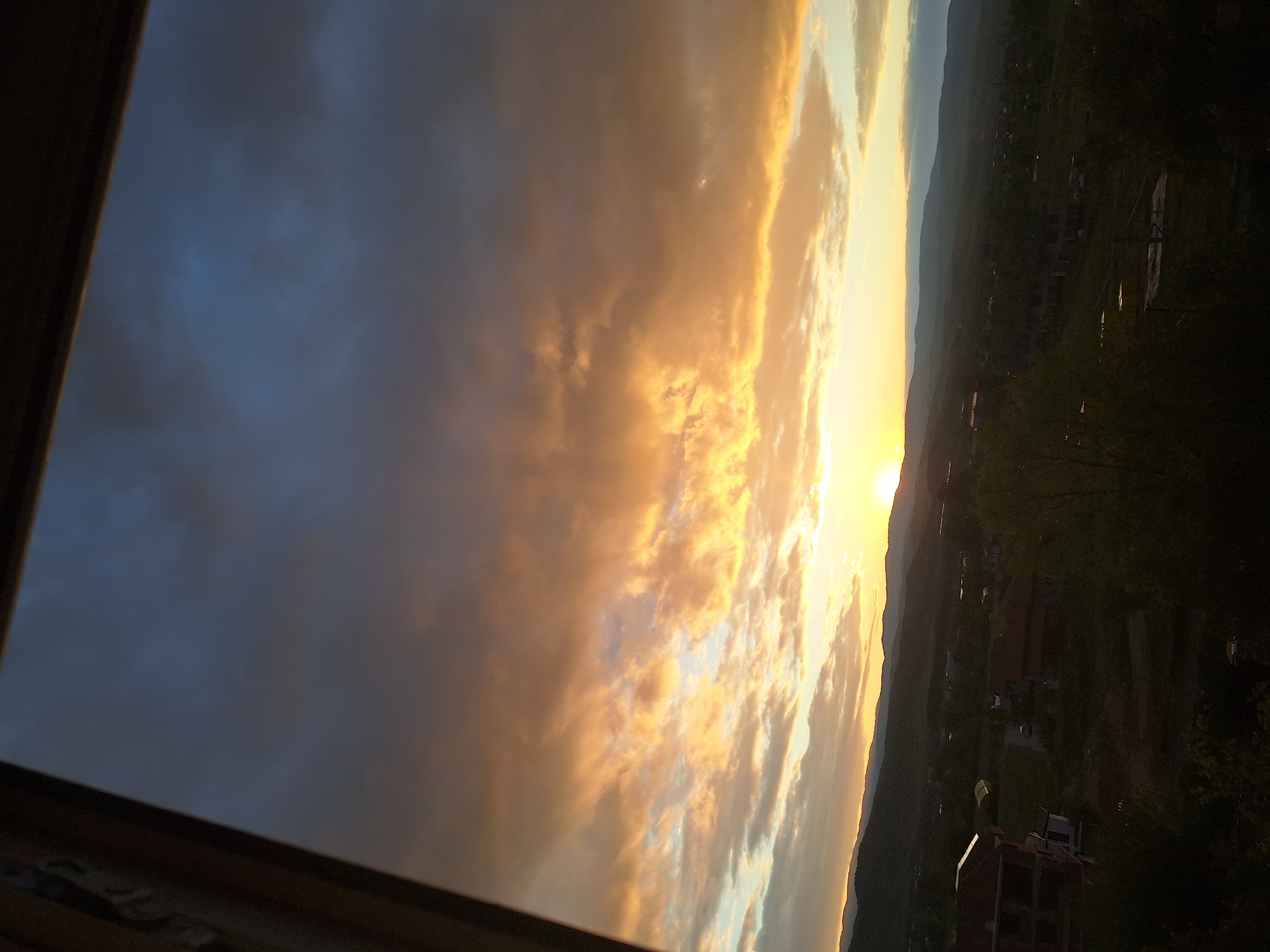 The image depicts a dramatic sunset or sunrise over a landscape, viewed from what appears to be a window. The sky is the dominant feature, filled with a dynamic display of clouds illuminated by the low-lying sun. The sun, visible on the right side of the frame (when correctly oriented), casts intense golden, orange, and yellow hues across the clouds immediately surrounding it. These brightly lit clouds contrast sharply with darker, blue-grey cloud formations occupying the upper left and central portions of the sky.
Below the vibrant sky, a distant horizon line reveals a silhouette of mountains or hills stretching across the landscape. The foreground and midground, largely obscured by shadow, show a dark expanse of land with subtle indications of buildings and sparse vegetation, appearing almost as dark silhouettes against the brighter sky. The overall lighting suggests either dawn or dusk, with the warm colors strongly indicative of a sunset.
A dark window frame runs along the bottom and left edges of the photograph, partially framing the view and suggesting the image was captured from an indoor vantage point looking outwards. The weather appears to be partly cloudy. No people or visible text are present in the image. The scene conveys a sense of natural beauty and tranquility, observed over what is stated to be Blagoevgrad, Bulgaria.
