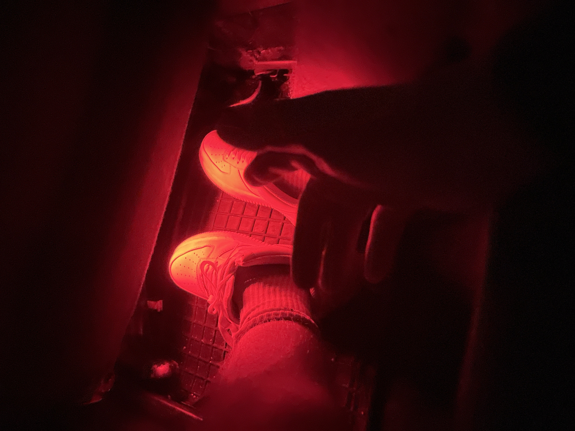 The image shows a close-up view of a person's feet inside a vehicle, illuminated by red interior lighting. The feet are clad in white sneakers with red laces, resting on a black textured floor mat. The toes of the right shoe are clearly visible, as are the laces and the sole. The left foot is partially obscured by the right leg and a dark shadow. The ambient lighting casts a strong red hue over the scene, creating a dramatic and somewhat mysterious atmosphere. The background is dark and out of focus, suggesting the interior of a car, possibly at night. The scene appears to be within a car's footwell.