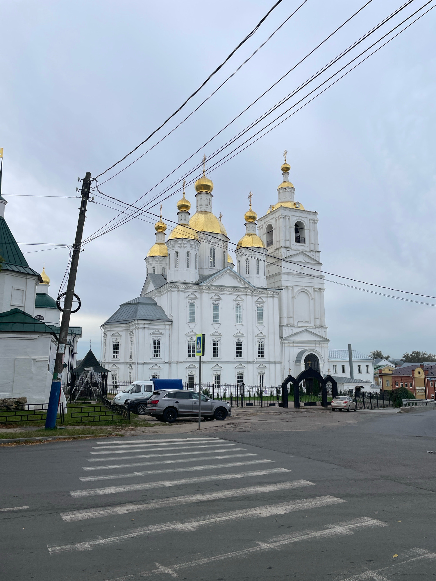 The image captures a frontal view of a large, white Orthodox cathedral with golden domes, situated in the city of Arzamas, Russia. The cathedral is prominently featured in the background, with its ornate architecture, including multiple domed structures and a bell tower, clearly visible against a cloudy sky. In the foreground, a pedestrian crossing is marked on an asphalt road. Two cars are parked near the cathedral entrance, one a grey SUV and another a lighter-colored sedan. A utility pole with numerous wires is positioned on the left side of the image, obscuring part of the cathedral. The scene appears to be a public space with a fence separating the road from the cathedral grounds.