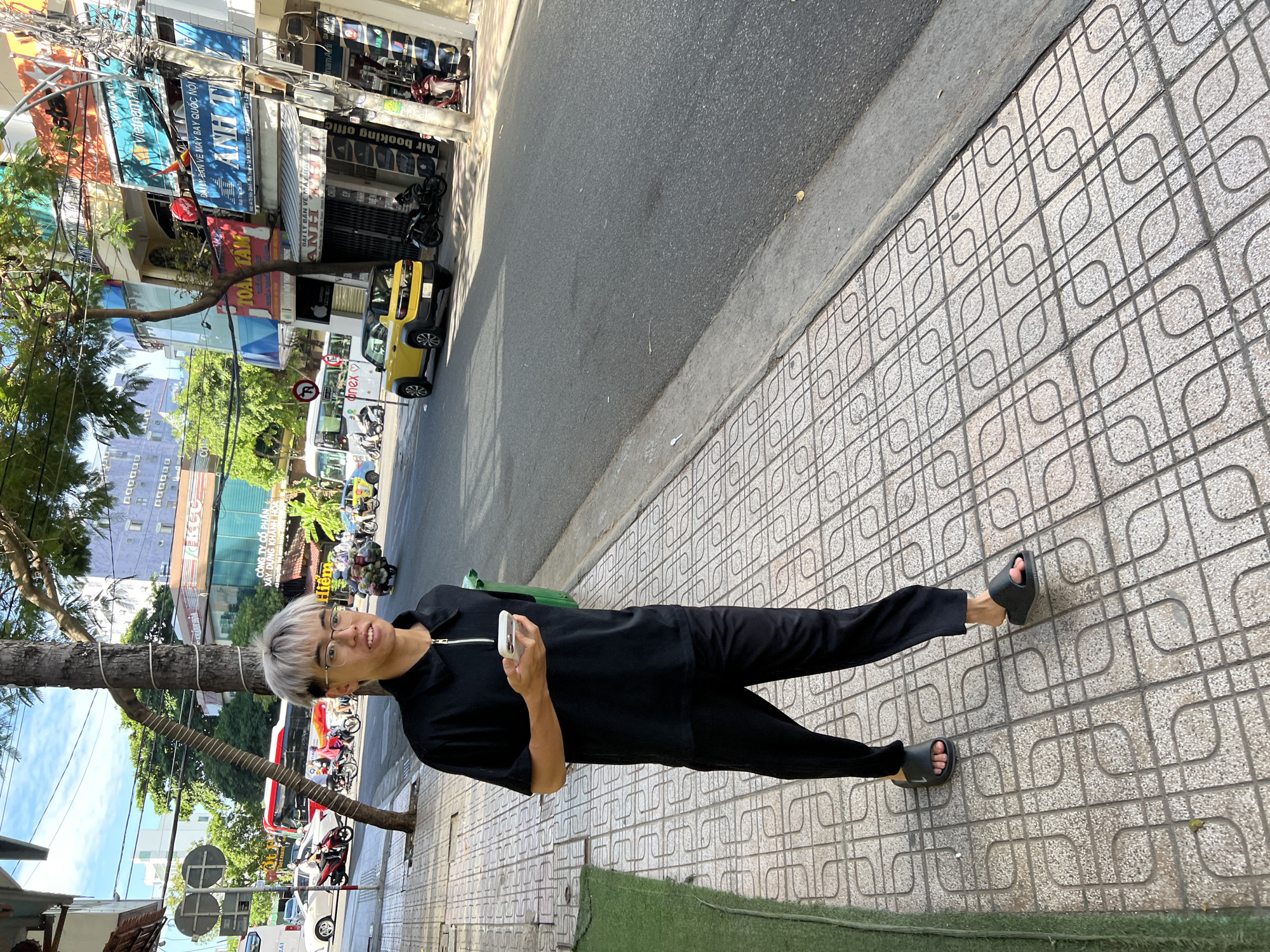 The image captures a street scene in Nha Trang, Vietnam. A young man with dyed blonde hair and glasses, wearing a black polo shirt with a zipper and black pants, walks on a tiled sidewalk. He is holding a white smartphone in his right hand. To his right, a green trash bin is visible.
The sidewalk has a patterned tile design. Adjacent to the sidewalk is a paved street with moderate traffic, including several motorcycles, a yellow car, and a white bus in the background. Buildings line the street, displaying various signs and advertisements in Vietnamese, indicating businesses such as "VietnamAir," "ANH T," and others. Trees with green foliage are present along the street and between buildings, suggesting a tropical urban environment. The sky is partly cloudy and bright, indicating daytime.