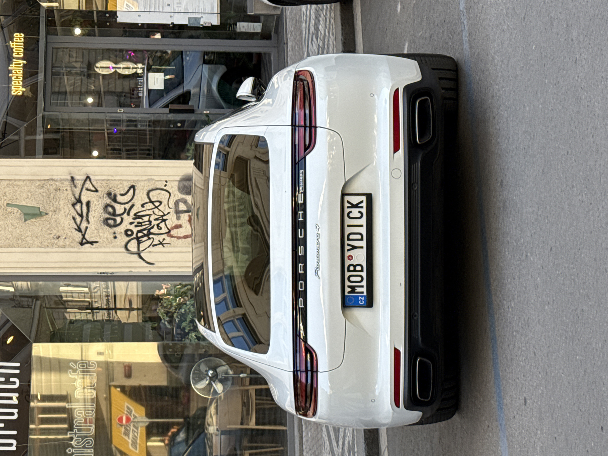 A white Porsche Panamera 4 is parked on a street in Prague, Czechia. The car is facing the camera and is positioned centrally in the frame. The license plate reads "MOB Y DICK" with "CZ" indicating it is from the Czech Republic. Behind the car, there is a building with a glass storefront and graffiti on the wall. The storefront has signs for "mistral café" and "specialty coffee". In the reflection of the glass, a street scene with buildings and parked vehicles can be seen. There are no people clearly visible in the image. The street appears to be paved and there are some faded blue lines on the ground, suggesting it is a parking area. The lighting indicates it is daytime, with sunlight casting reflections on the car and building.