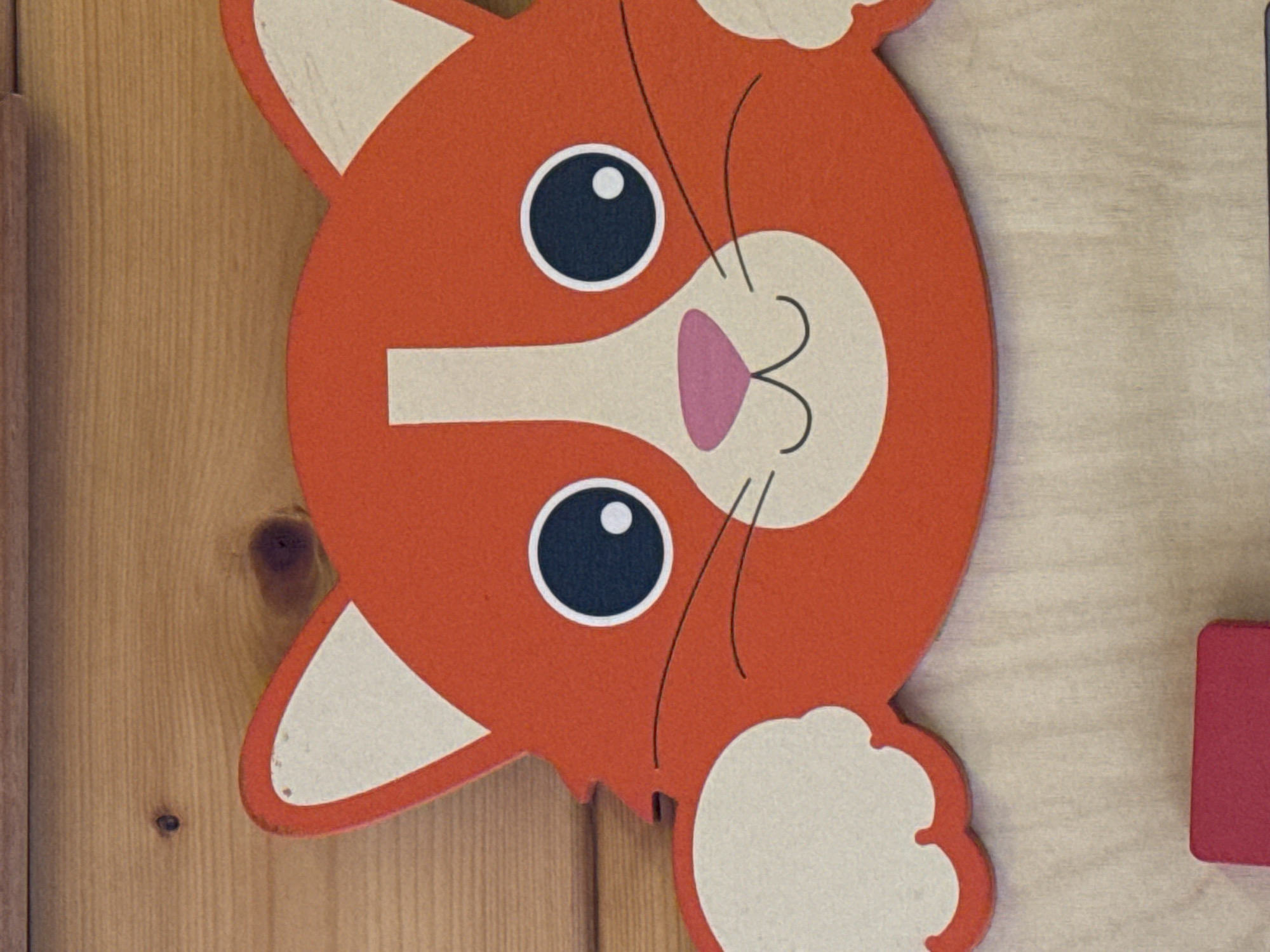 A close-up shot features a stylized, cartoonish cat's head, predominantly orange-red, set against a wooden background. The cat's head is inverted within the frame, with its pointed ears directed towards the bottom. It displays large black circular eyes with white pupils, a light beige muzzle, and a small pink heart-shaped nose. Delicate black lines depict whiskers on either side of the muzzle. The cat's inner ears and what appear to be fluffy cheek or paw details are also rendered in the light beige color. The background is a light-toned wooden surface, exhibiting distinct vertical grain patterns and a few darker wood knots, suggesting an indoor environment. A small, solid red rectangular block is partially visible along the right edge of the image. No activity is taking place, and there is no visible text. The image presents a static scene of these objects. While the location context is Ново-Никольское, Russia, no specific visual elements in the image confirm this.