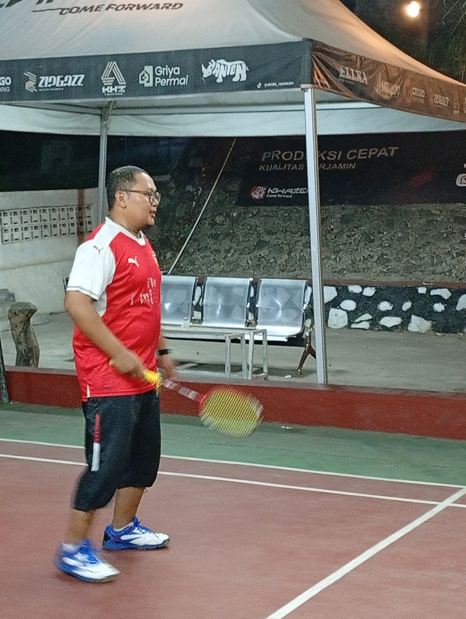 A medium shot captures a man playing badminton on an outdoor court in Caringin, Indonesia. The man, wearing glasses, a red and white polo shirt, and black shorts, is mid-action, holding a yellow badminton racket. His left foot is blurred due to motion. The court is red with green sections and white lines. In the background, a white tent with various logos and text is set up, with several empty metal chairs beneath it. Behind the tent, a stone-patterned wall is visible. The lighting suggests it is evening or night.