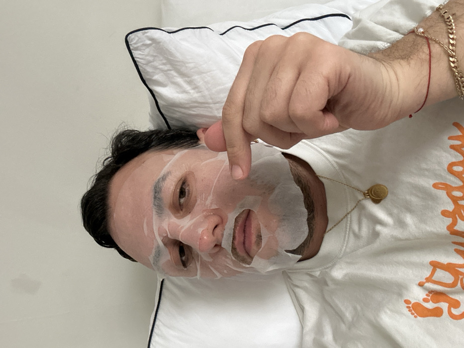 An adult male is depicted in a close-up, selfie-style photograph, lying down with his head resting on a white pillow trimmed with navy blue piping. His face is covered by a translucent sheet mask, with cutouts for his partially open eyes, nostrils, and mouth. Dark hair is visible above his forehead. He displays stubble or a short beard beneath the mask around his chin and upper lip. He wears a white crew-neck t-shirt featuring orange text, partially visible as "Thursday" alongside a footprint graphic. A gold-toned locket-style pendant on a thin chain hangs around his neck. His left hand is raised, forming a 'finger heart' gesture with the tips of his thumb and index finger touching. A thin red string bracelet and a gold-colored chain bracelet adorn his left wrist. The background is a plain white wall, exhibiting a faint, light-colored stain in the upper left quadrant. No visual information within the image definitively identifies a specific location within the city of London, United Kingdom.