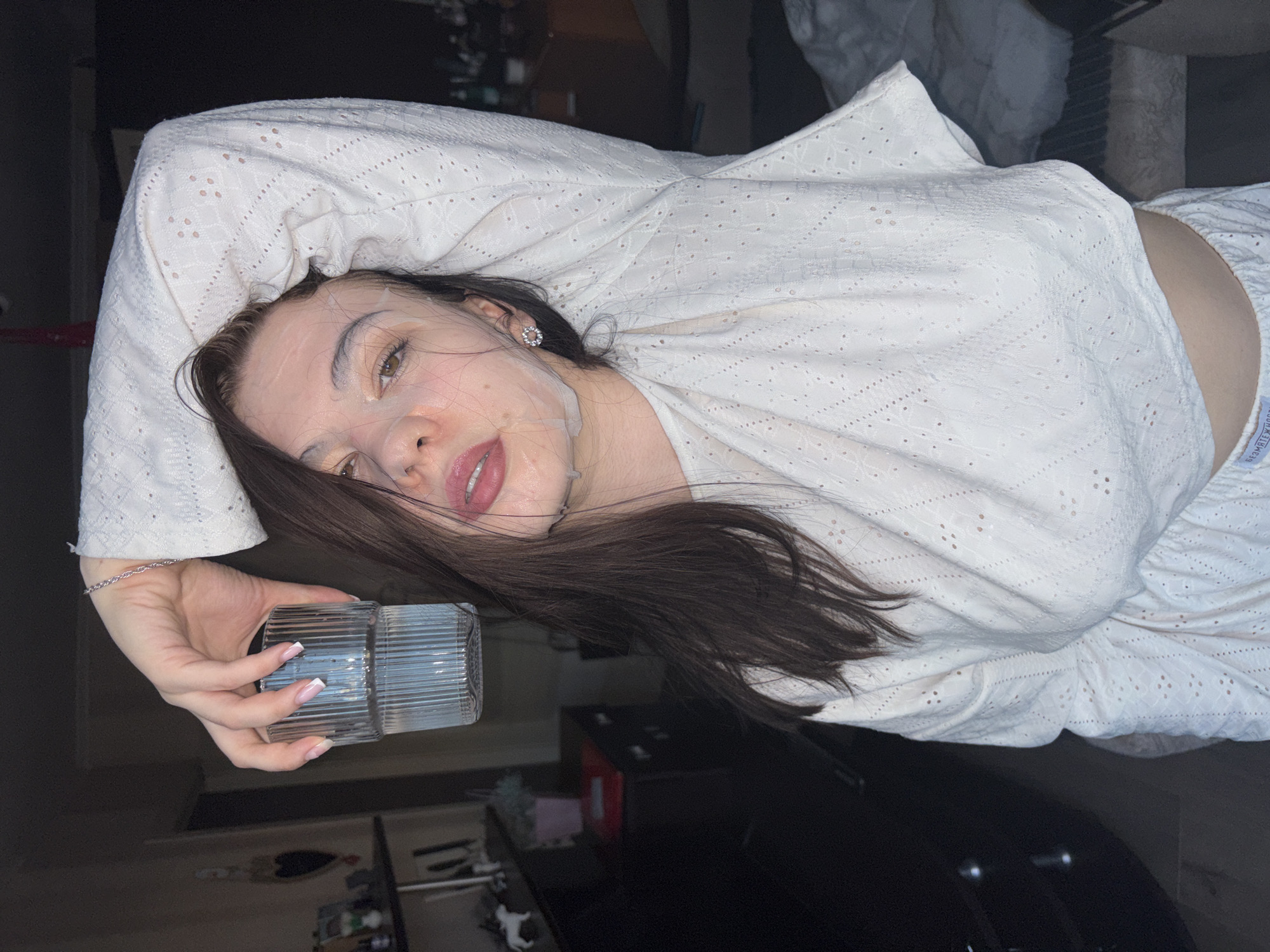 The scene depicts a young adult female located indoors in an unidentifiable residential setting within Perm, Russia. The subject has long, dark brown hair and is wearing a translucent sheet face mask, which appears moist on her skin. Her eyes are light brown, and her lips are slightly parted, showing a pink hue. She is wearing a two-piece white garment made of eyelet fabric with small perforations, consisting of a long-sleeved top and what appears to be matching shorts; the waistband of the shorts bears the text "БЕЗМЯТЕЖНОСТЬ" (Serenity). A silver chain bracelet is visible on her left wrist, and she wears a small, circular, silvery earring in her right ear. Her fingernails have a white tip (French manicure style).
The female is positioned with her left arm raised and bent at the elbow, holding a clear, ribbed glass containing water. The glass is held above her head, slightly to the left of the frame's center. Her right arm is bent, with the hand near her head, partially obscured by her hair. She looks directly towards the camera.
The background is dimly lit, revealing a dark shelf or cabinet unit on the left side of the frame. On this unit, various objects are discernable: a white figurine (possibly a horse or dog), several vertical decorative items, and a small plant in a pink pot. A dark, heart-shaped wall hanging is visible further back on the left. To the right of the subject, a rumpled blanket or duvet is partially visible, suggesting a bed or sofa. The overall lighting is low, with the subject illuminated from the front, casting the background into shadow.