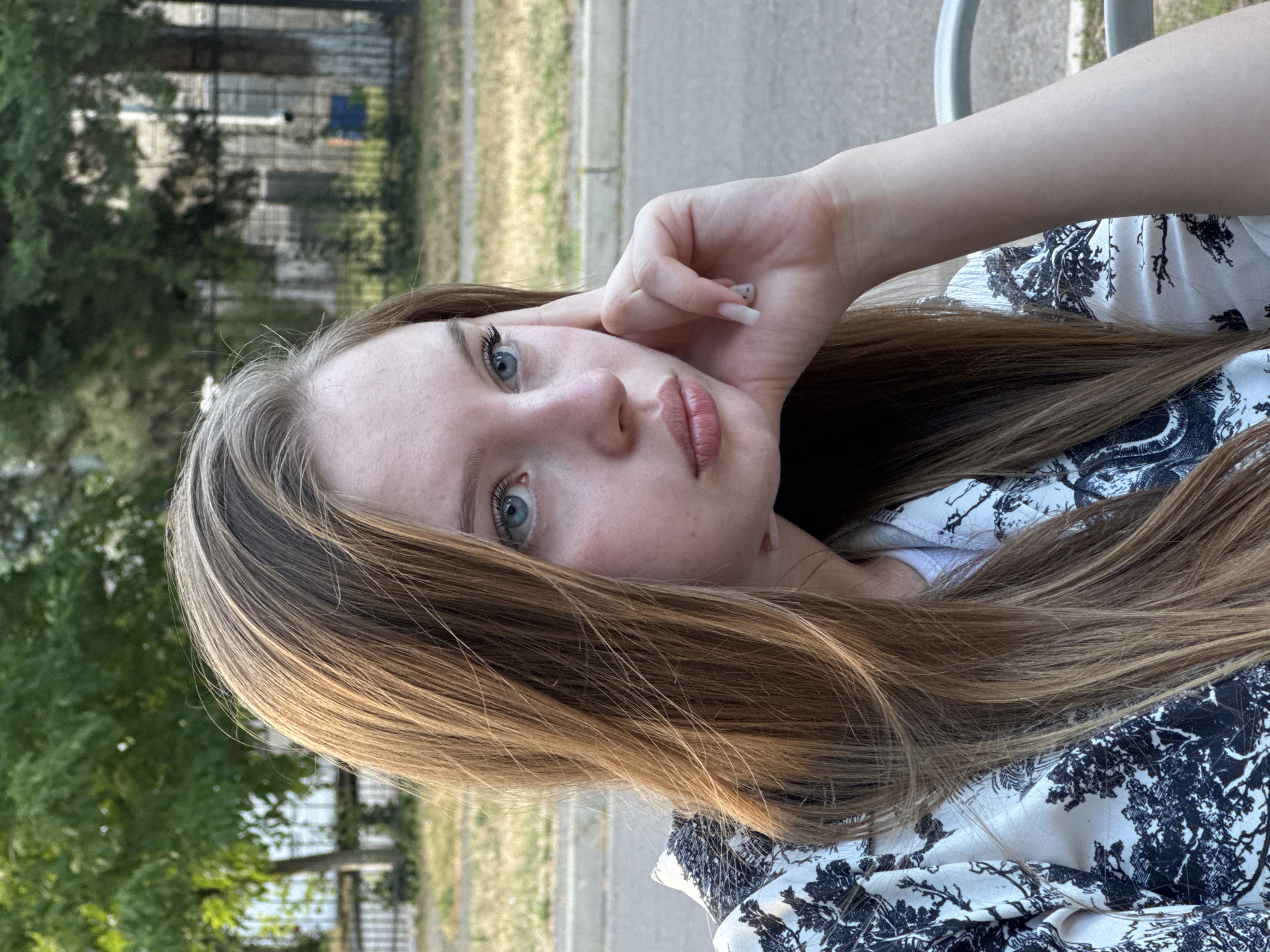 A close-up, sideways shot focuses on the face of a young woman with long, light brown hair. Her hand is raised, touching her forehead and temple, with her fingers resting near her eye. Her eyes are light blue, and she has light-colored skin and full lips with a subtle sheen. She is wearing a white top with a black pattern. The background is blurred, showing trees and a fence, suggesting an outdoor setting, likely in Tiraspol, Moldova. The lighting indicates it is daytime, possibly late afternoon, given the warm tones. There is no visible text in the image.