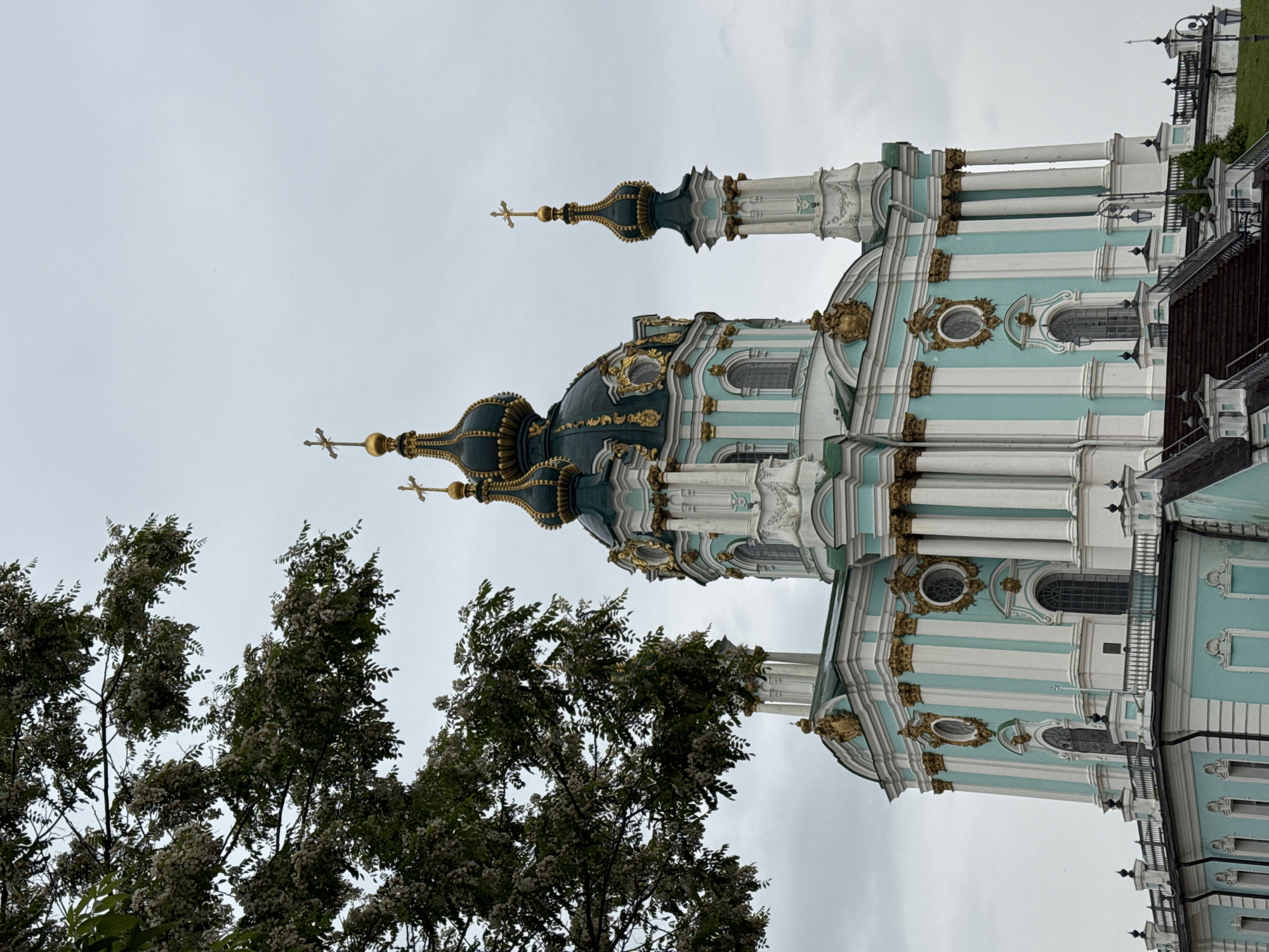 This media file captures a detailed view of St. Andrew's Church in Kyiv, Ukraine, under an overcast sky. The prominent subject is the ornate Baroque-style church, characterized by its distinctive light teal and white striped exterior, richly adorned with gold decorative elements and stucco work. Multiple domes, topped with golden onion-shaped cupolas and Orthodox crosses, are visible, including a larger central dome and smaller surrounding ones.
The church's facade showcases columns, arched windows, and intricate architectural details typical of the Rococo period. The lighting is soft and even due to the cloudy conditions, indicating daytime. In the foreground, partially obscuring the lower-left portion of the church, are trees with lush green foliage and numerous small white blossoms, suggesting springtime or early summer. No people or visible text are present in the image. The scene conveys a sense of architectural grandeur and historical significance within an outdoor setting.