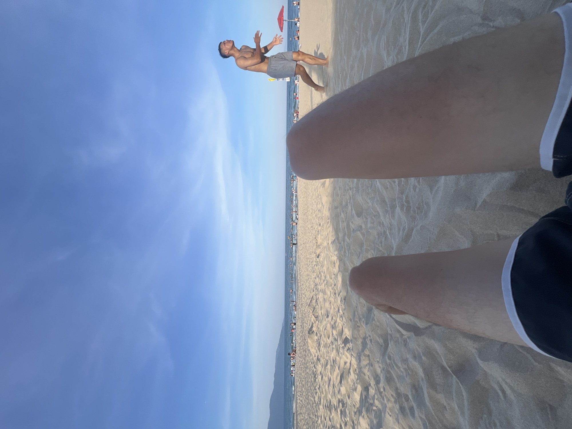 The image shows a first-person perspective of someone relaxing on a sandy beach in Đà Nẵng, Vietnam. The foreground is dominated by the subject's legs and thighs, with sandy dunes stretching out behind them. In the mid-ground, the ocean meets the shore, with a clear blue sky overhead. A line of people are visible along the water's edge, some swimming and others relaxing on the sand. In the background, mountains rise in the distance, forming a scenic backdrop. A young man in grey shorts is in the process of playing a game or sport on the sand, his body angled and his arms raised in preparation for an action. A red umbrella stands in the sand to his right. The overall scene depicts a sunny day at the beach, with a mix of recreational activities and natural beauty.