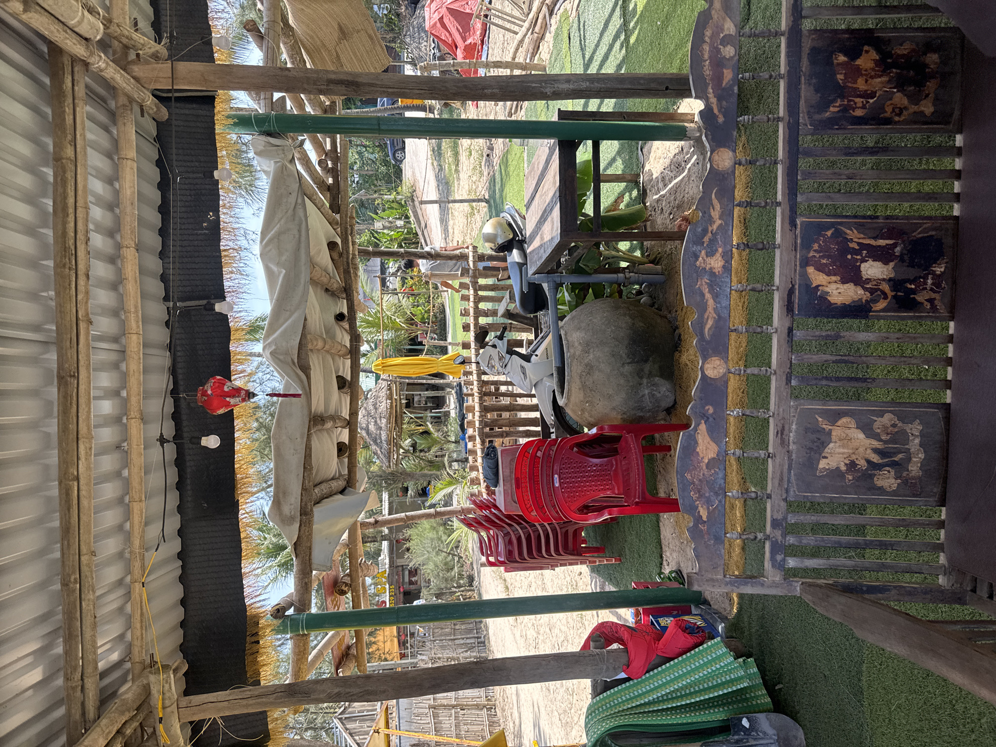 The image depicts an outdoor seating area in Đà Nẵng, Vietnam. The scene features a variety of objects including red plastic chairs, a stack of green mats and red items, a stone, a table with a helmet on it, and a scooter in the background. A decorative wooden screen is also visible. The area is partially shaded by bamboo structures and thatched roofs, and a string of lights is hanging. In the distance, buildings, vegetation, and people can be seen.