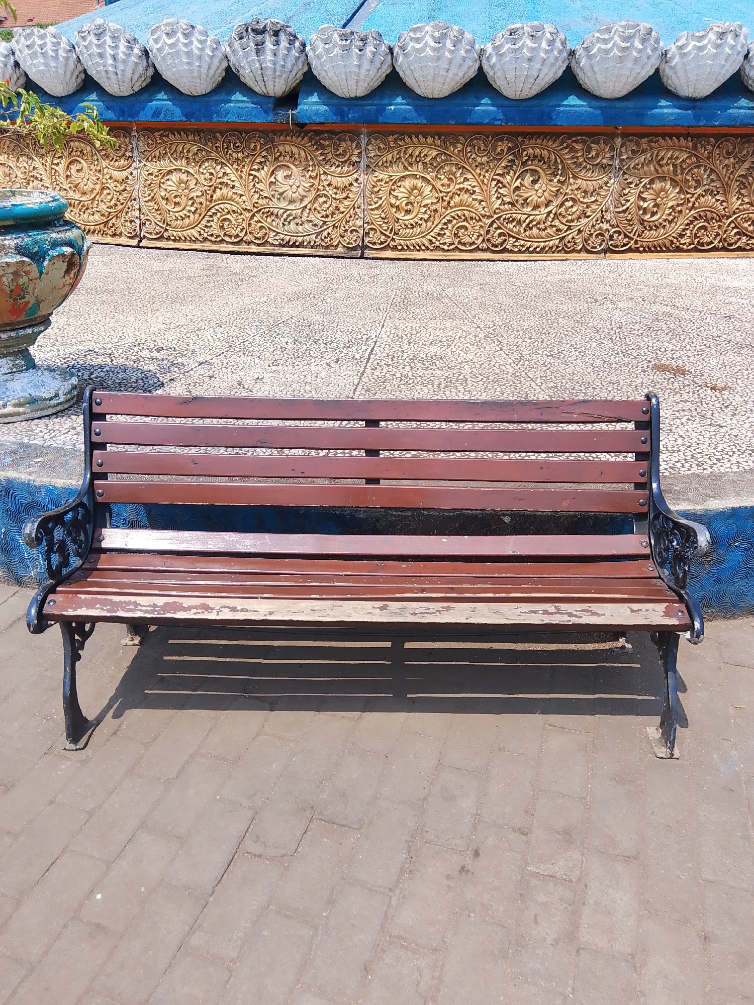 A weathered, dark red wooden bench with black wrought iron legs sits on a paved area in front of an ornate golden wall featuring intricate floral carvings. Above the wall, a blue band is adorned with a row of decorative, shell-like white ornaments. To the left of the bench, a portion of a blue and green patterned planter is visible. The scene is illuminated by direct sunlight, casting shadows from the bench onto the paving stones. The overall setting suggests an outdoor public space, likely in a location such as Tahunan, Indonesia, given the architectural style and the presence of decorative elements.