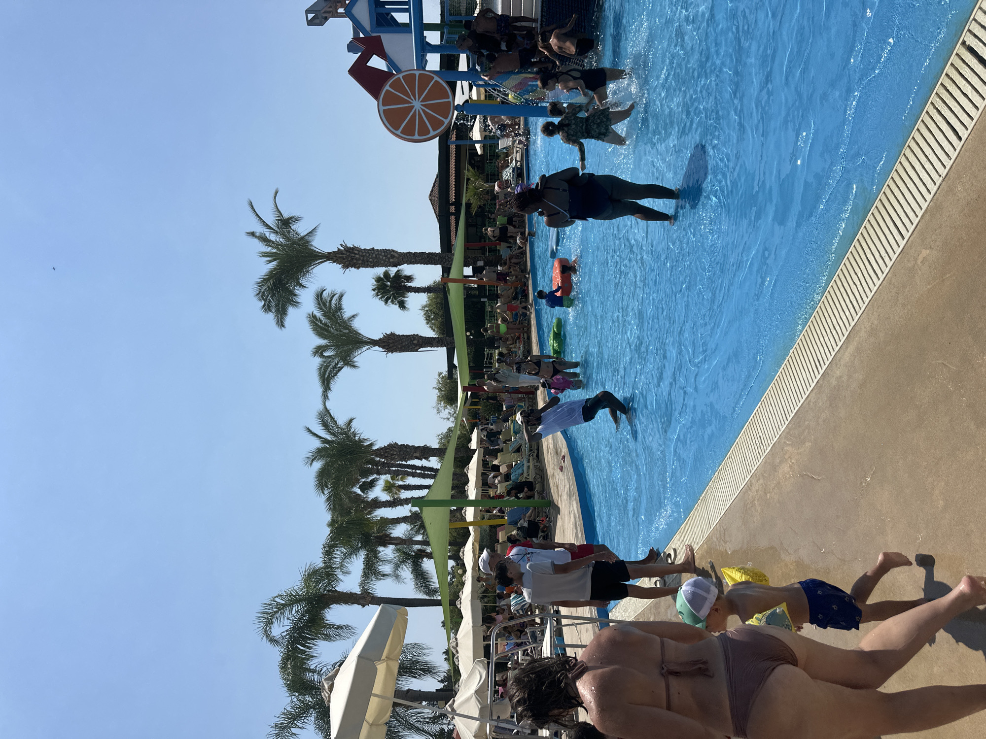 This image depicts a sunny day at a crowded outdoor swimming pool, possibly a water park, located in an unknown city. Numerous people are engaged in various water activities, including swimming, wading, and playing on inflatable toys. Palm trees are visible in the background, suggesting a warm climate. A large pool with clear blue water dominates the scene, with a tiled edge separating it from a paved walkway. Several lounge chairs and umbrellas are set up around the pool area, indicating a recreational setting. A water slide structure with colorful components is also present. The sky is a clear blue, with no clouds visible.