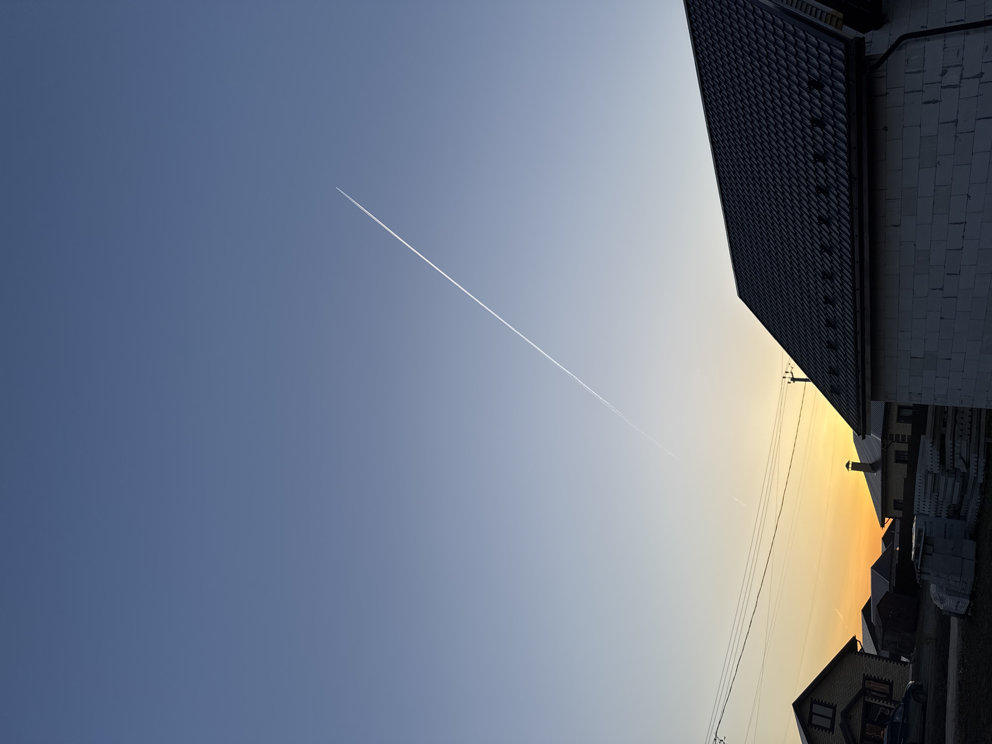 The image shows a view of the sky in Gornyi, Russia, at sunset. The sky transitions from blue to yellow-orange. A contrail from an airplane cuts across the sky. Parts of buildings, including roofs and walls constructed from bricks or blocks, are visible at the edges of the image, suggesting a residential area. Overhead power lines are also visible against the sky.